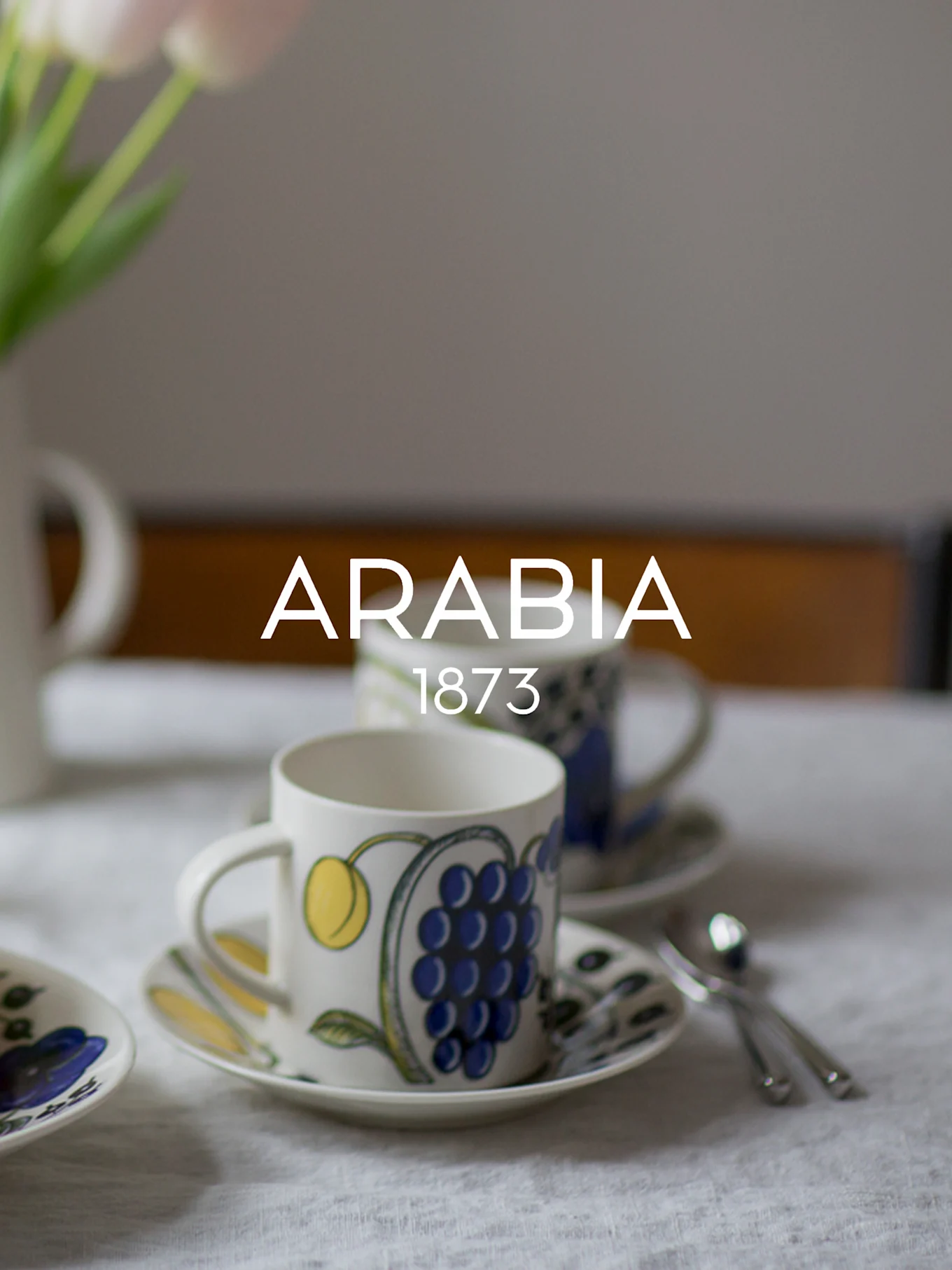 Due tazze e piattini in ceramica decorati, uno con uva blu, su una tovaglia bianca. Il testo 'ARABIA 1873' è sovrapposto.
