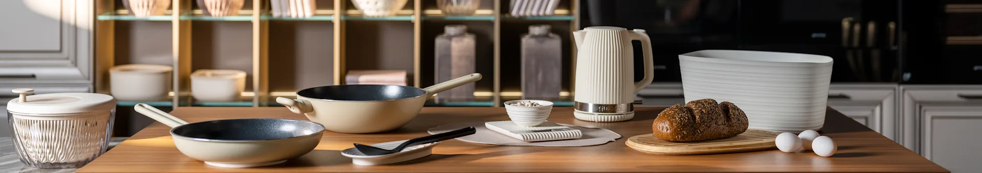 Bancone da cucina con padelle beige, un bollitore elettrico bianco a coste, una pagnotta di pane ai semi su un tagliere e uova. Accessori da cucina moderni.