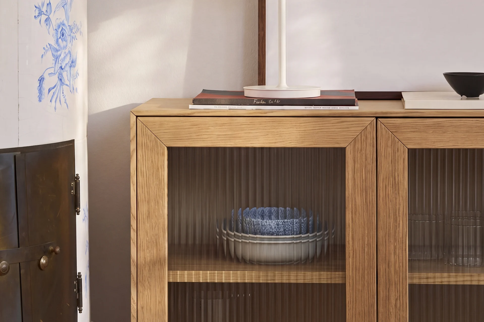 Un mobile in legno chiaro con ante in vetro scanalato contiene ciotole blu e bianche, accanto a una stufa in ceramica con motivi floreali blu.