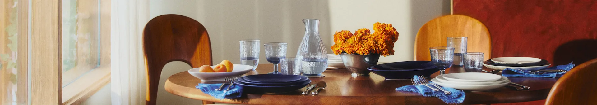 Tavolo da pranzo rotondo in legno con piatti blu e bianchi, tovaglioli blu e fiori arancioni. La luce del sole filtra da una finestra.