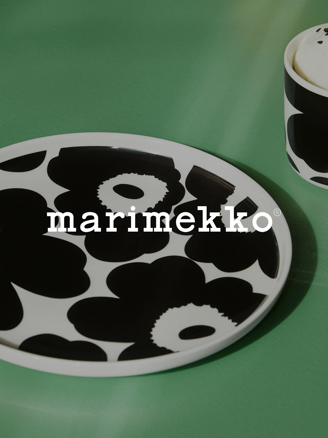 Piatto ovale bianco con motivo floreale nero e logo Marimekko su una superficie verde, con una tazza abbinata parzialmente visibile.