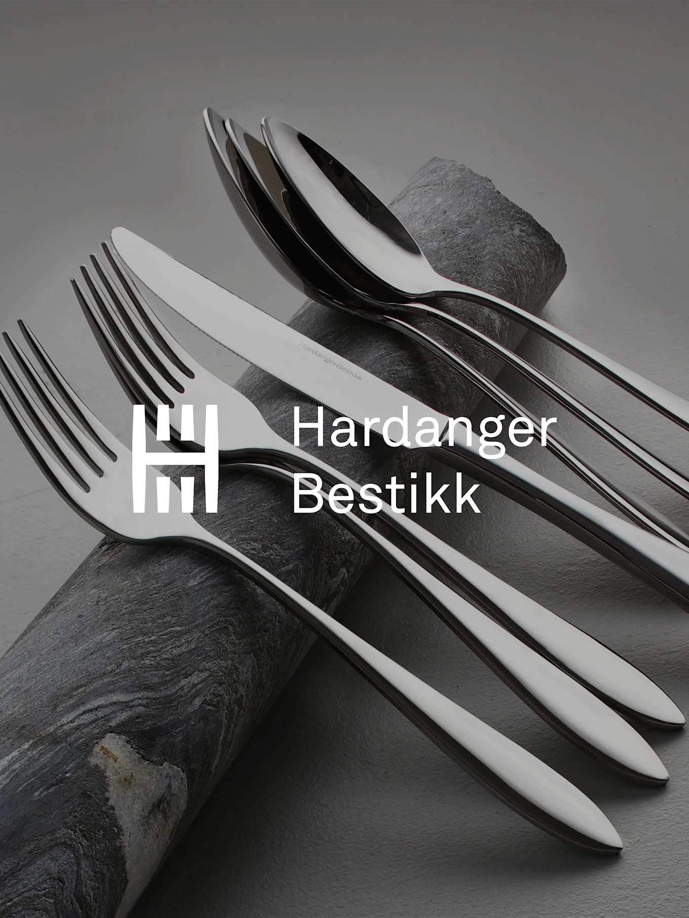 Set di posate in acciaio inossidabile lucido, incluse forchette, coltelli e cucchiai, artisticamente disposte su pietra scura e texturizzata.