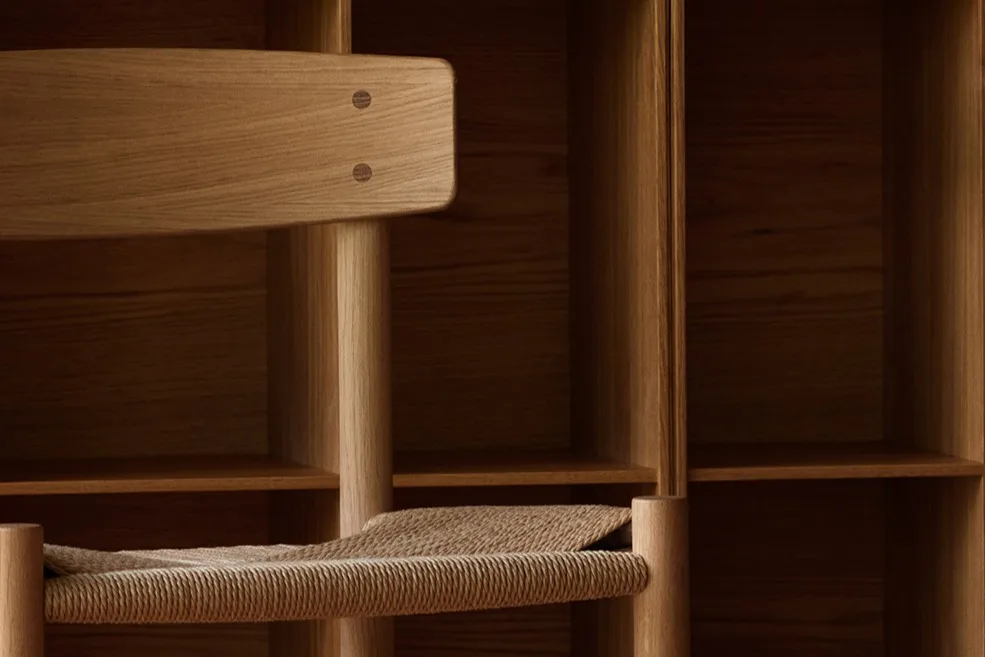 Una sedia in legno chiaro con seduta intrecciata davanti a una parete di scaffali quadrati in legno e un pavimento in legno a motivi.
