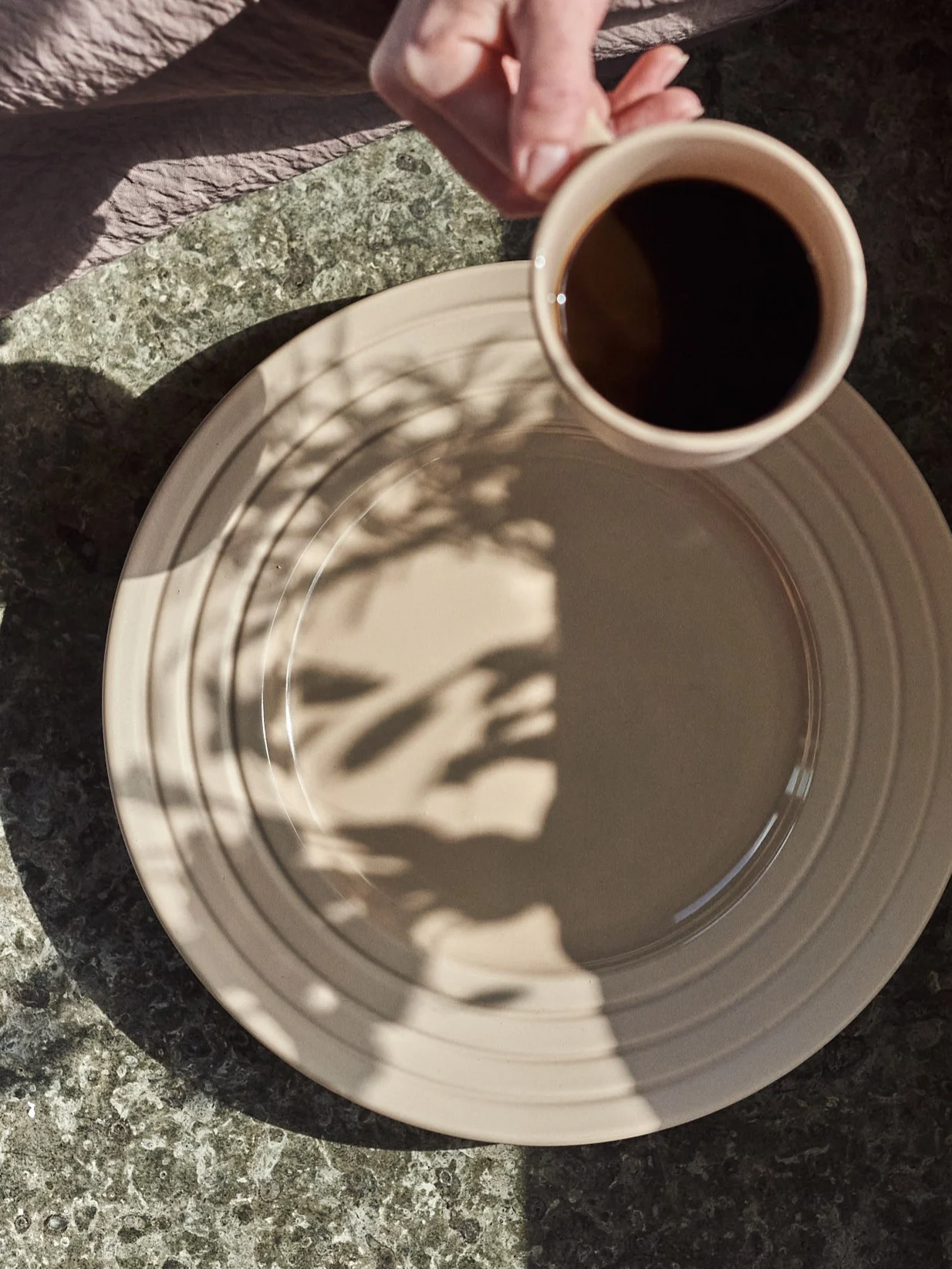 Mano tiene tazza di caffè sopra piatto beige con ombre di foglie su un tavolo maculato.