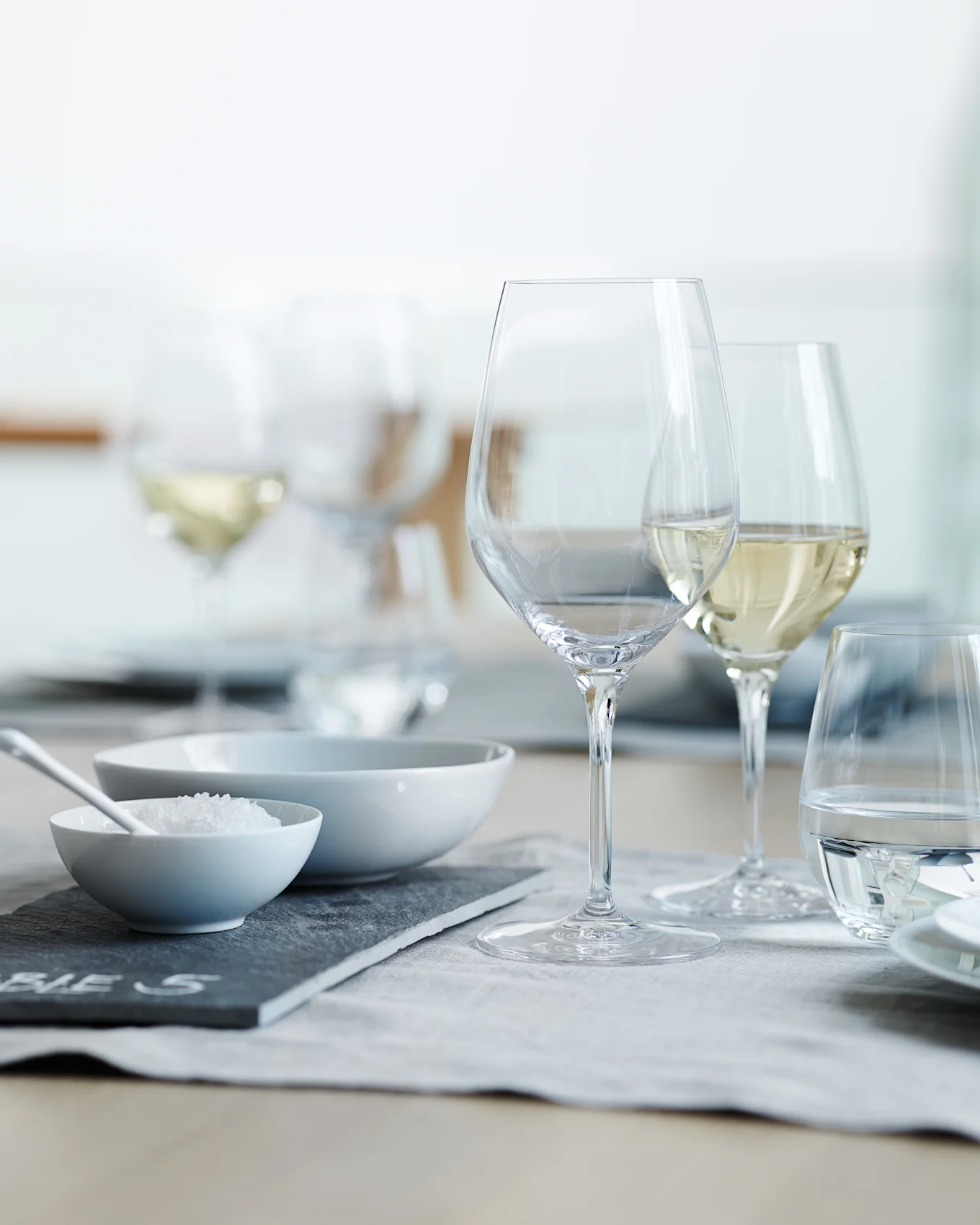 Una tavola apparecchiata con bicchieri da vino e acqua trasparenti, una ciotola di sale grosso e una ciotola vuota su una tovaglietta grigia.