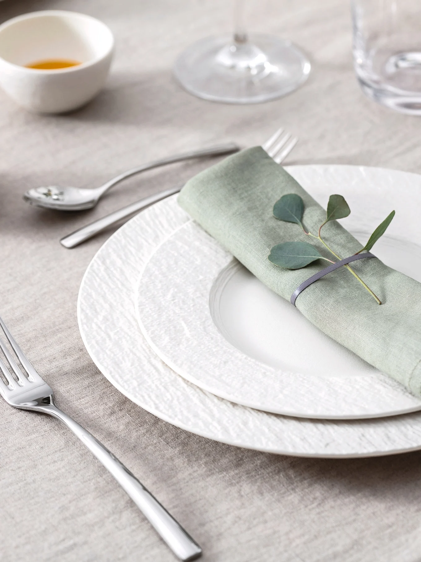 Elegante mise en place con tovagliolo di lino verde chiaro, foglie di eucalipto, piatti bianchi testurizzati e posate su una tovaglia beige.