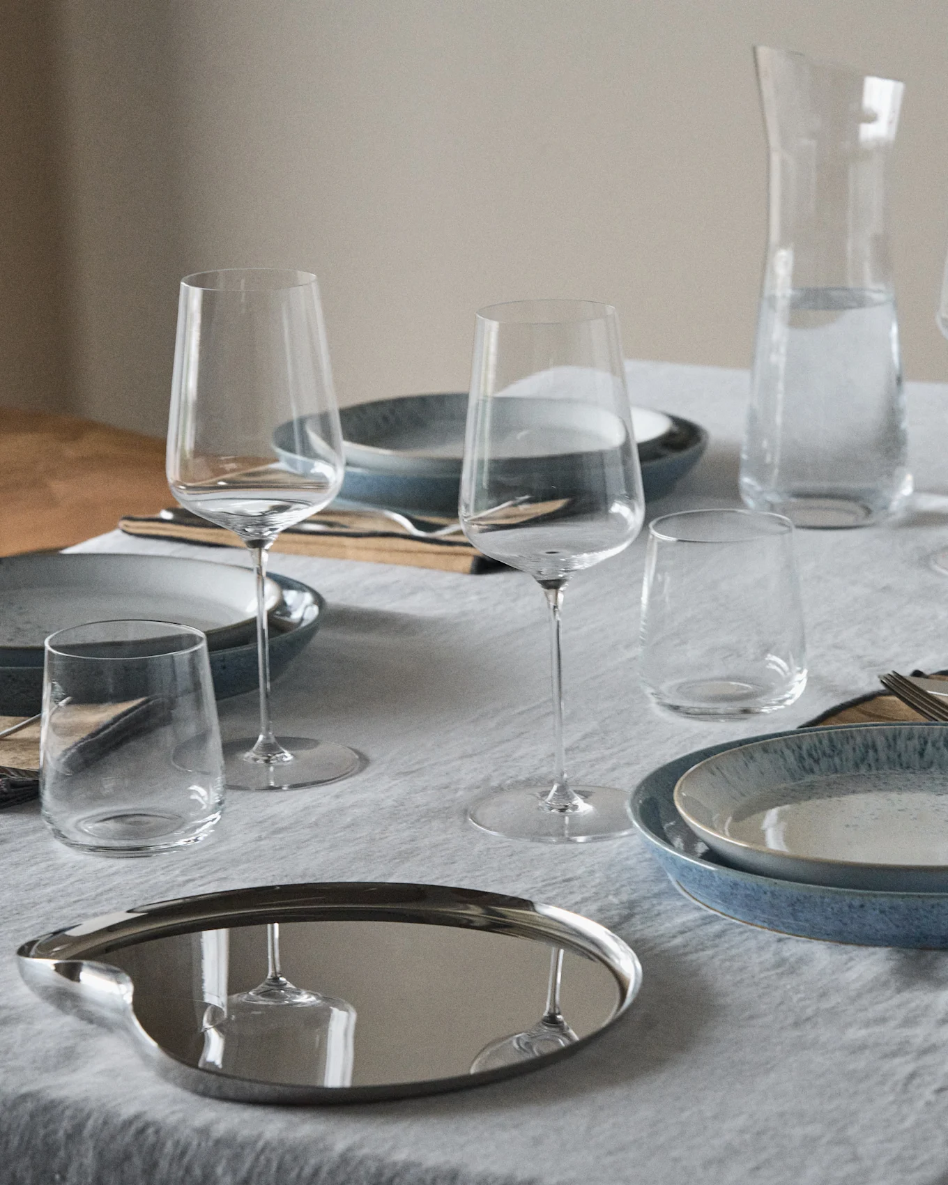 Tavolo apparecchiato con piatti blu e maculati, calici da vino, bicchieri, una caraffa d'acqua e un vassoio ovale riflettente.