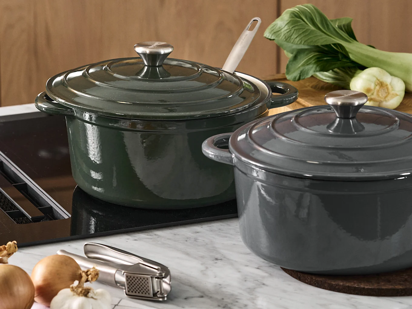 Due pentole in ghisa con coperchio, una verde scuro e una grigio scuro, su un piano di lavoro della cucina con bok choy, cipolle e uno spremiaglio.