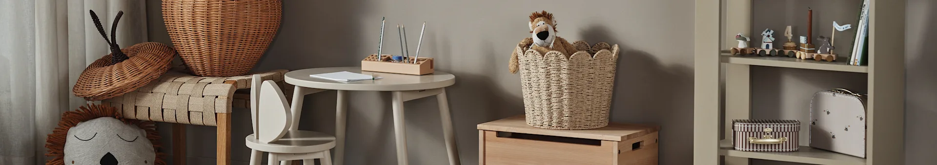 Una cameretta con un peluche a forma di leone in un cesto intrecciato, un tavolino di legno con matite e una libreria con giocattoli e libri.
