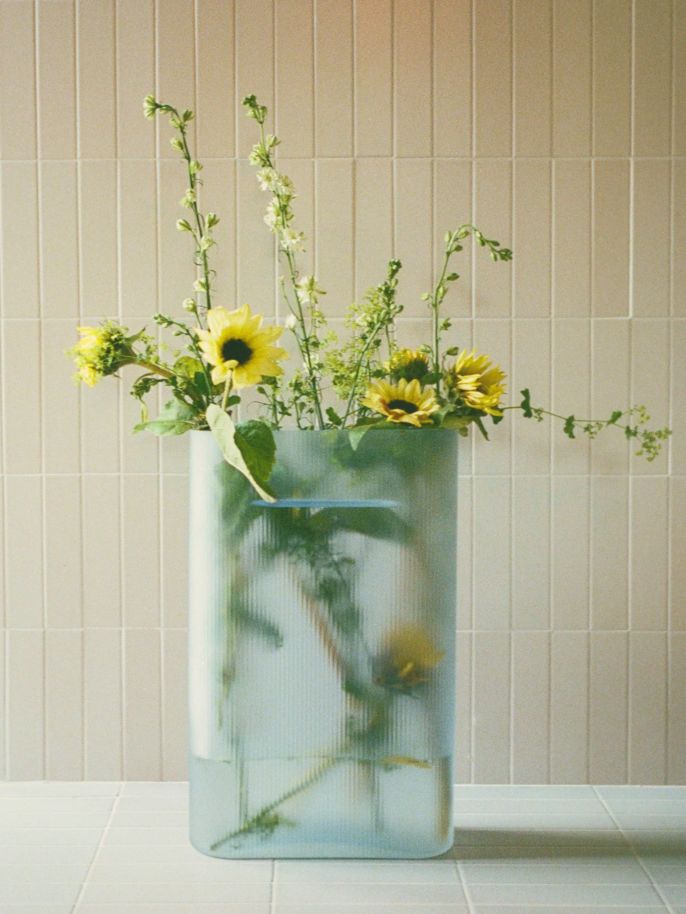 Mazzo di girasoli gialli e fiori verdi appuntiti in un vaso rettangolare trasparente e a coste su una superficie piastrellata.