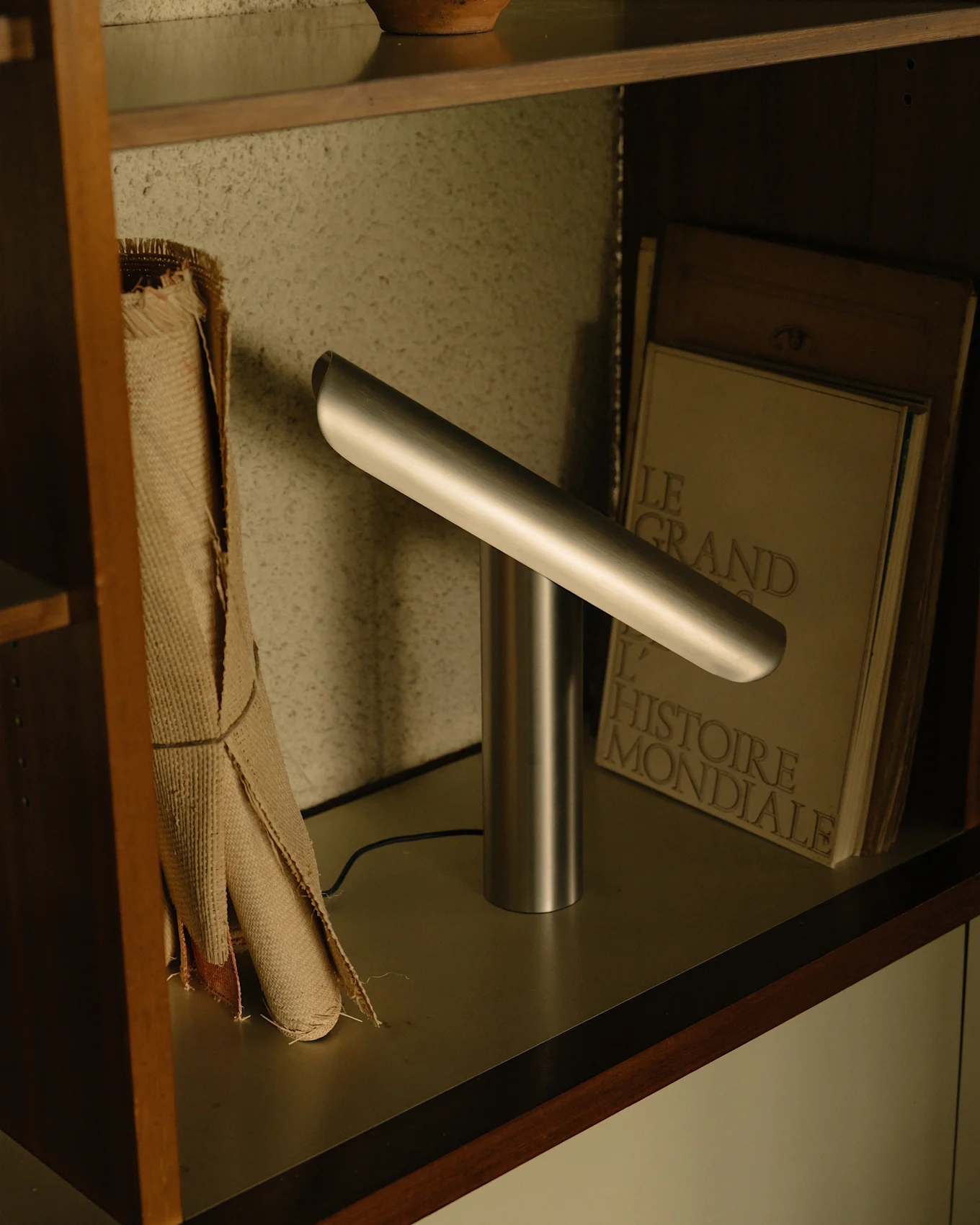 Lampada da scrivania moderna argentata con base cilindrica e barra luminosa orizzontale angolata su uno scaffale.