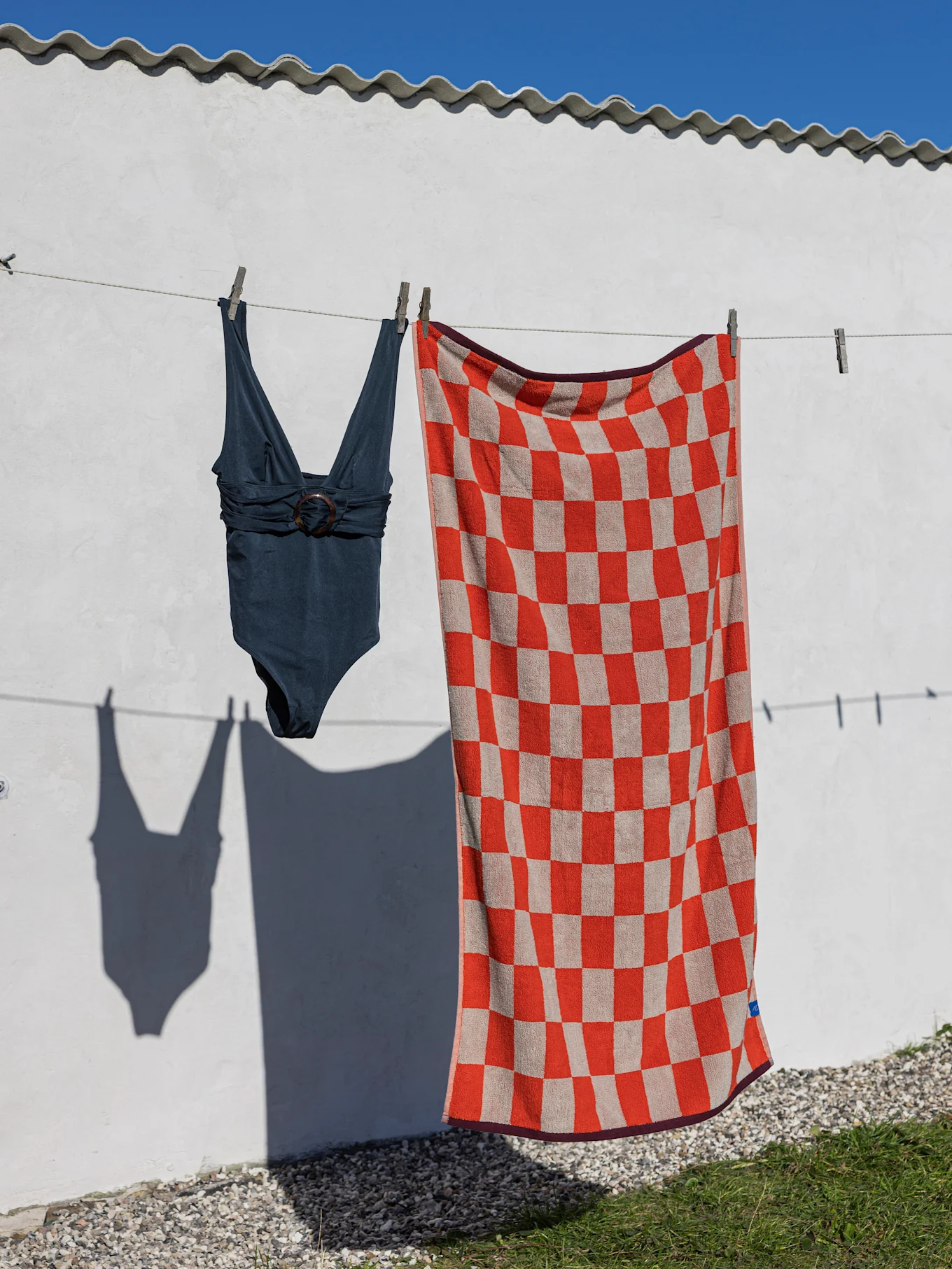 Un costume da bagno intero blu scuro e un telo mare a scacchi rossi e beige appesi a uno stendibiancheria su una parete bianca.