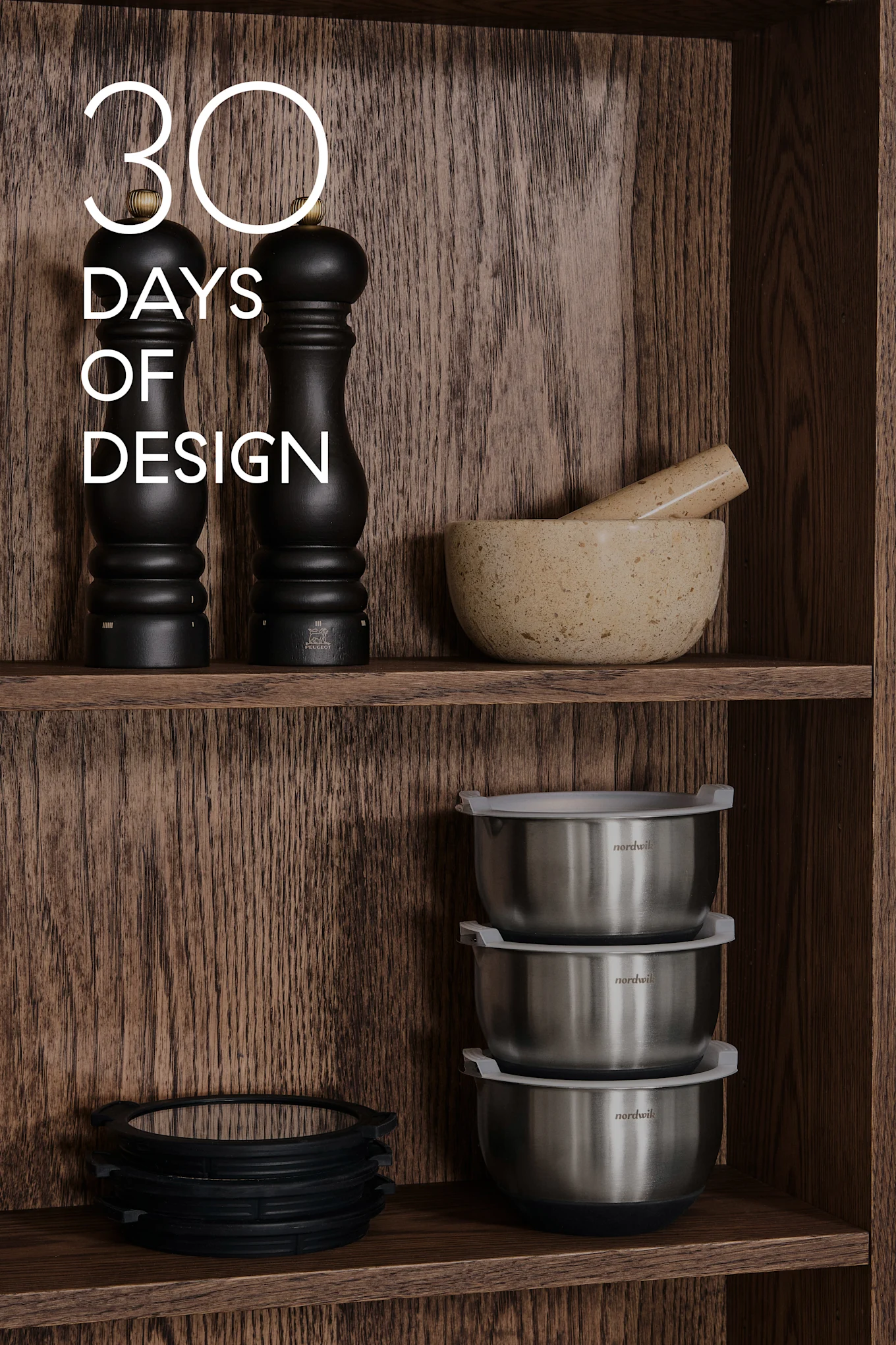 Scaffali in legno con utensili da cucina: macinini neri, mortaio e pestello, ciotole in acciaio, anelli in silicone. Testo: 30 DAYS OF DESIGN.