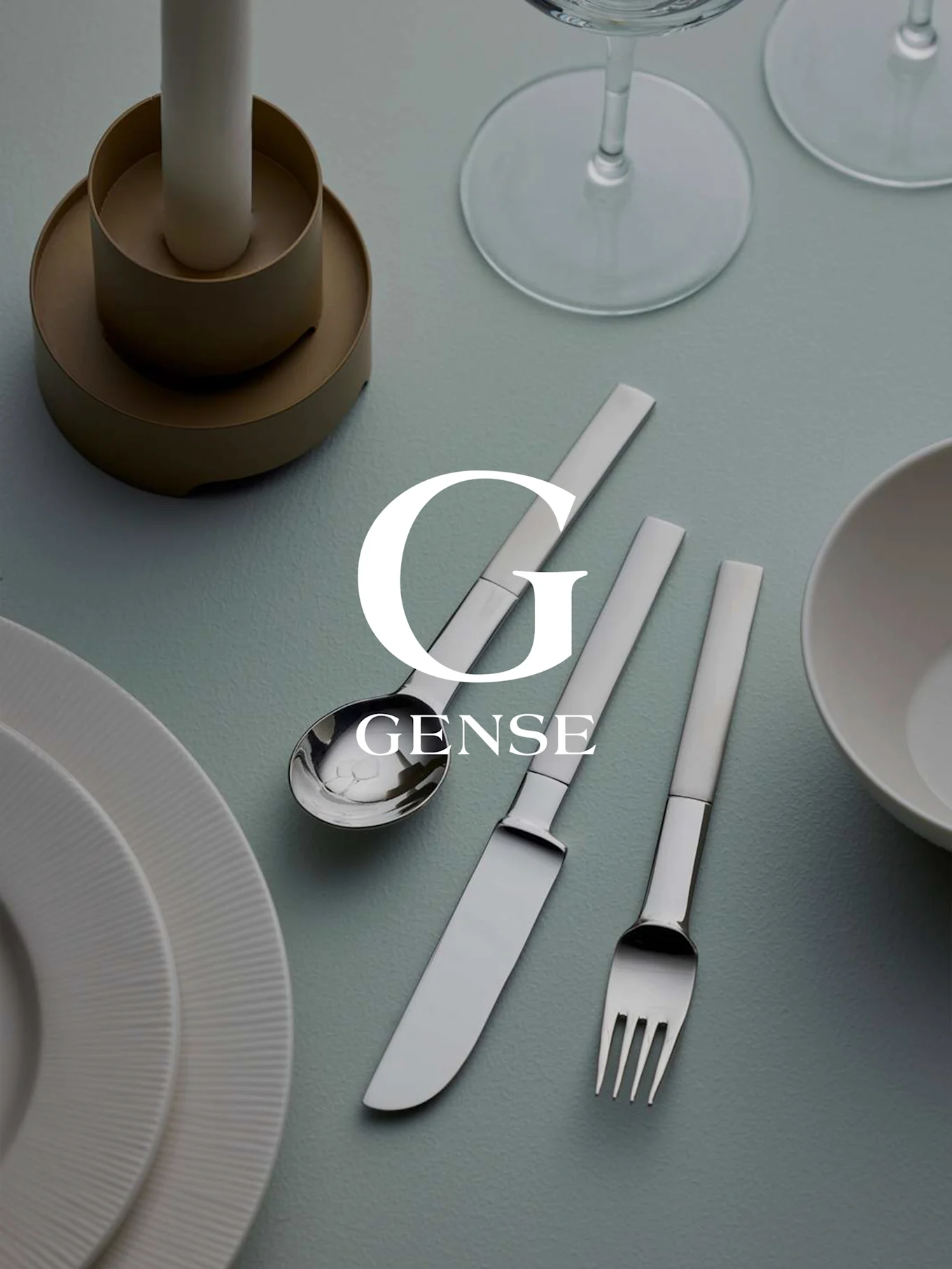 Una tavola apparecchiata con set di posate Gense in argento (cucchiaio, coltello, forchetta), piatti bianchi, una candela e bicchieri su superficie azzurra.