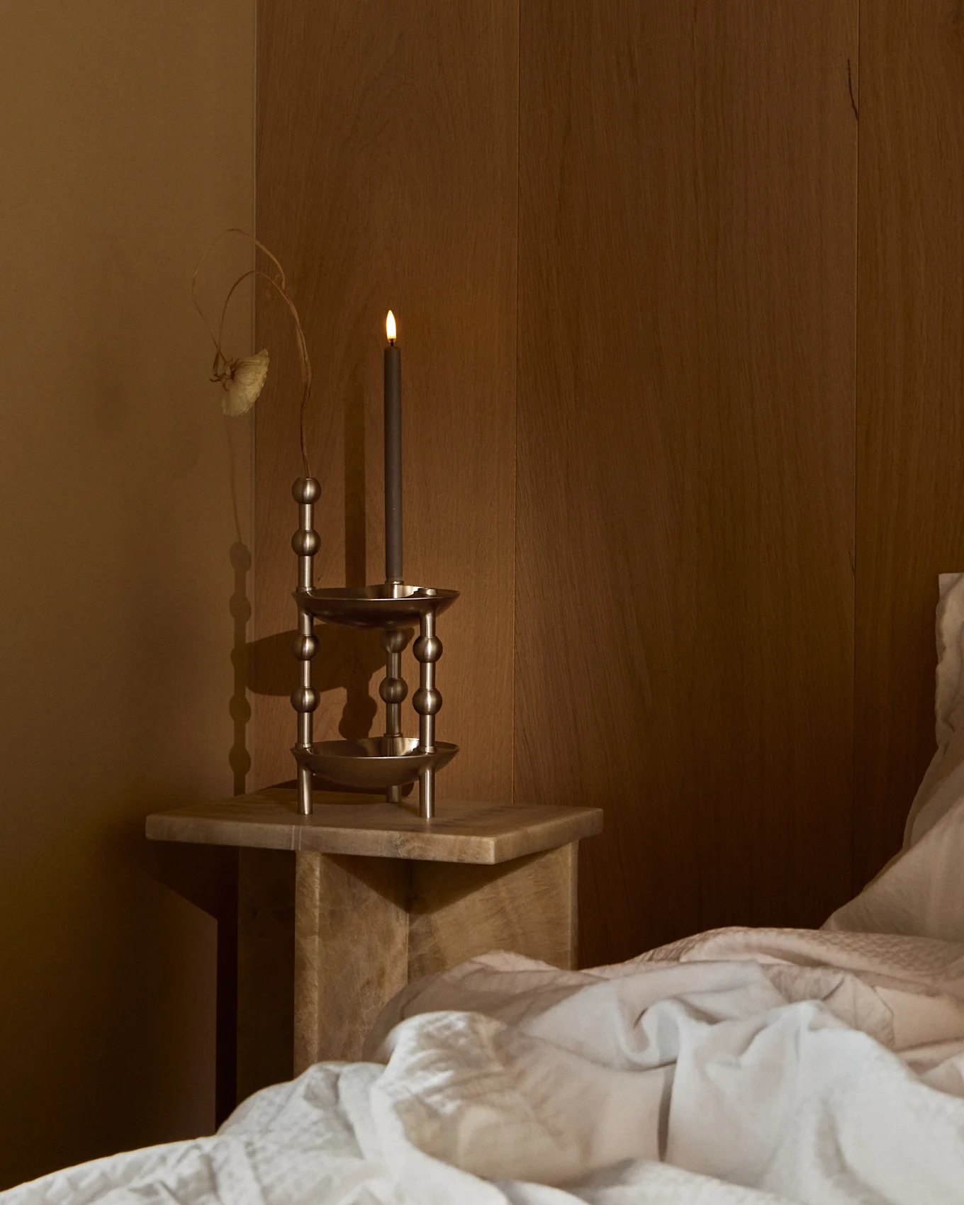 Una candela scura accesa in un portacandele metallico ornato su un comodino di legno accanto a un letto con biancheria da letto bianca in una stanza in penombra.