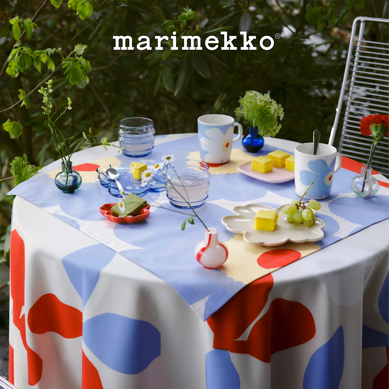 Tavolo esterno con tovaglia Marimekko a motivi rossi, blu, bianchi, apparecchiato con tazze floreali, dessert e piccole piante. Logo Marimekko.