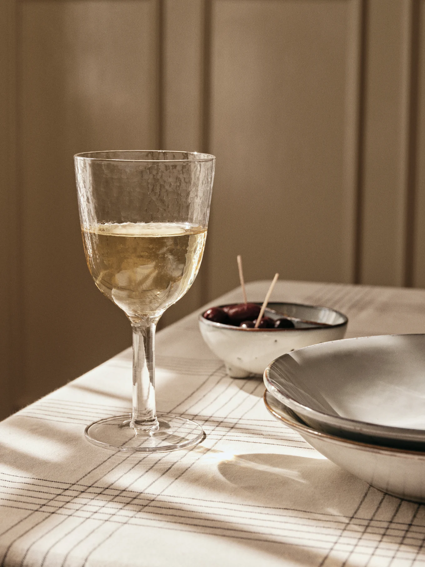 Un bicchiere di vino bianco strutturato su una tovaglia a righe, con olive e piatti sullo sfondo, immerso nella luce della finestra.