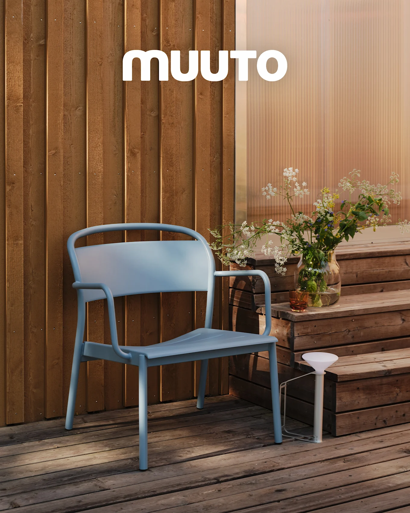 Una sedia da esterno azzurra su un ponte di legno accanto a gradini con un vaso di fiori selvatici e una lampada portatile bianca. Logo MUUTO.