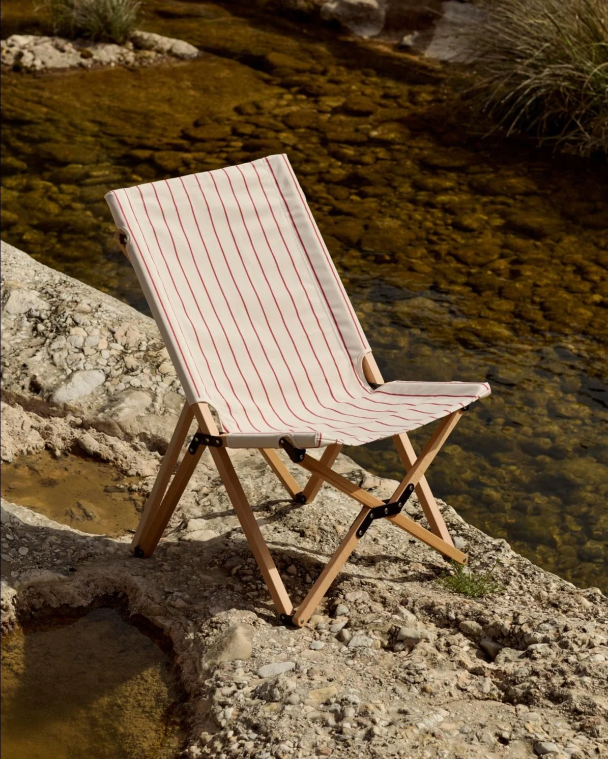 Sedia pieghevolmente con seduta in tessuto a righe bianche e rosse su una riva rocciosa accanto a un fiume.