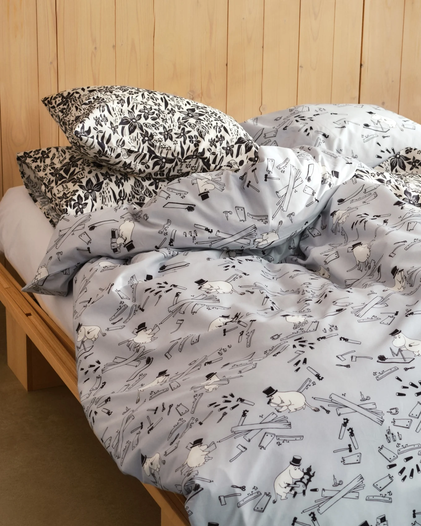 Letto con cuscini a stampa floreale bianca e nera e piumino azzurro con personaggi dei cartoni animati, attrezzi e assi di legno. Struttura del letto in legno.
