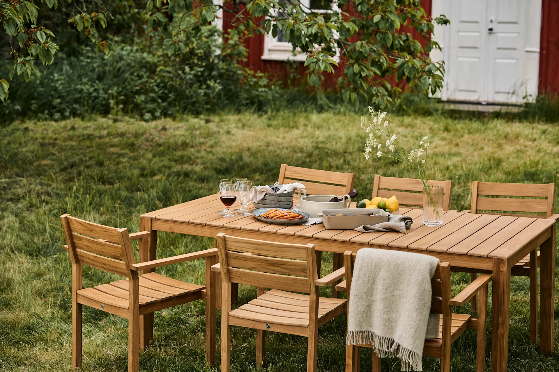 Set da pranzo esterno con tavolo in legno e sei sedie su un prato, apparecchiato con cibo, bevande e una coperta, con una casa rossa sullo sfondo.
