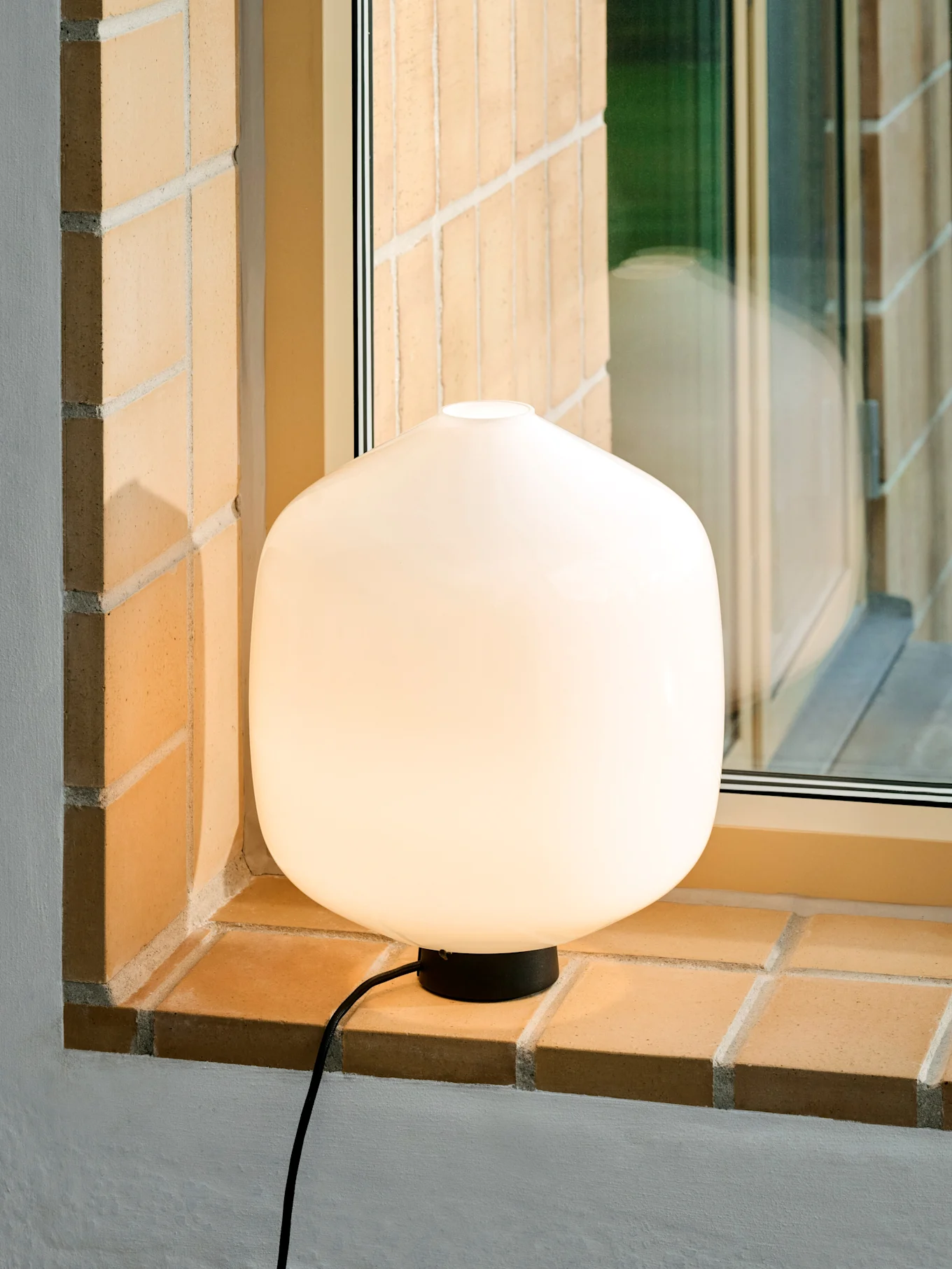 Una lampada da tavolo bianca, luminosa e arrotondata con base e cavo neri su un davanzale di mattoni.