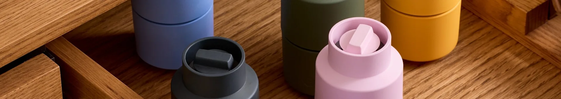 Cinque contenitori colorati: grigio scuro, azzurro, oliva, rosa chiaro con tappo unico e giallo senape, su legno.