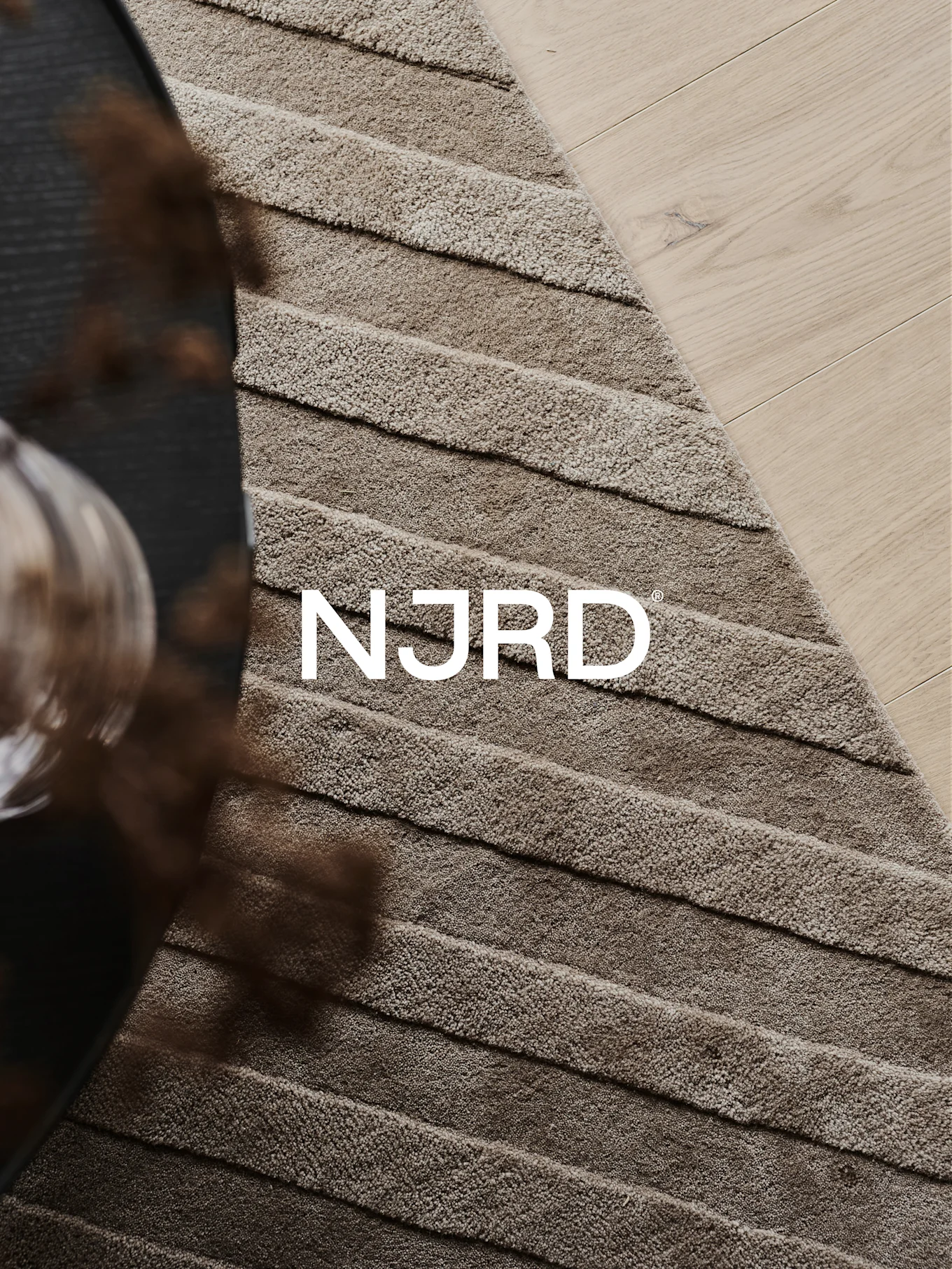 Primo piano di un tappeto shaggy marrone chiaro con creste orizzontali, parzialmente su un pavimento in legno chiaro, con il logo bianco 'NJRD'.