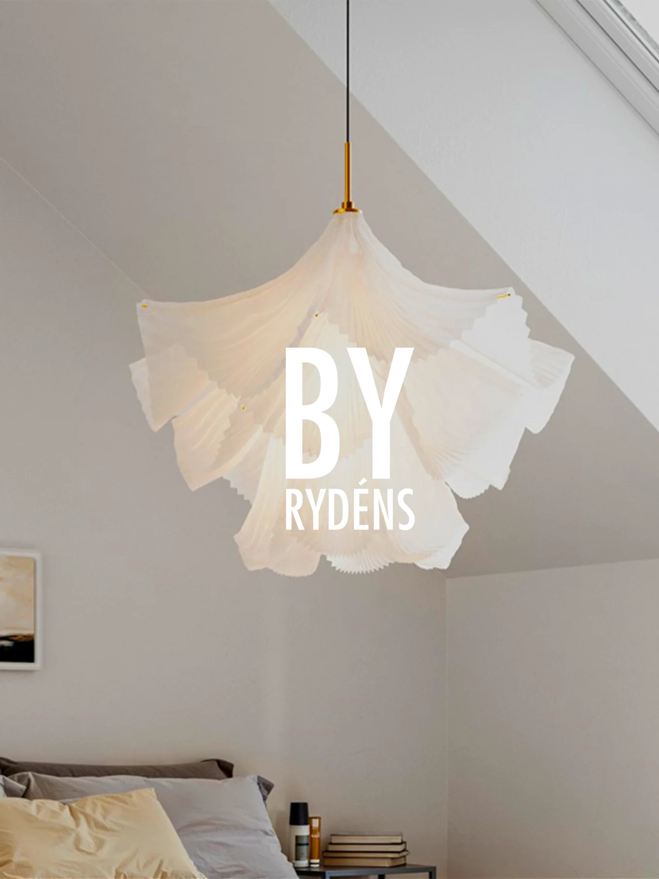 Una lampada a sospensione scultorea bianca e plissettata pende sopra un letto con cuscini e un comodino. 'BY RYDÉNS' è sovrapposto alla lampada.
