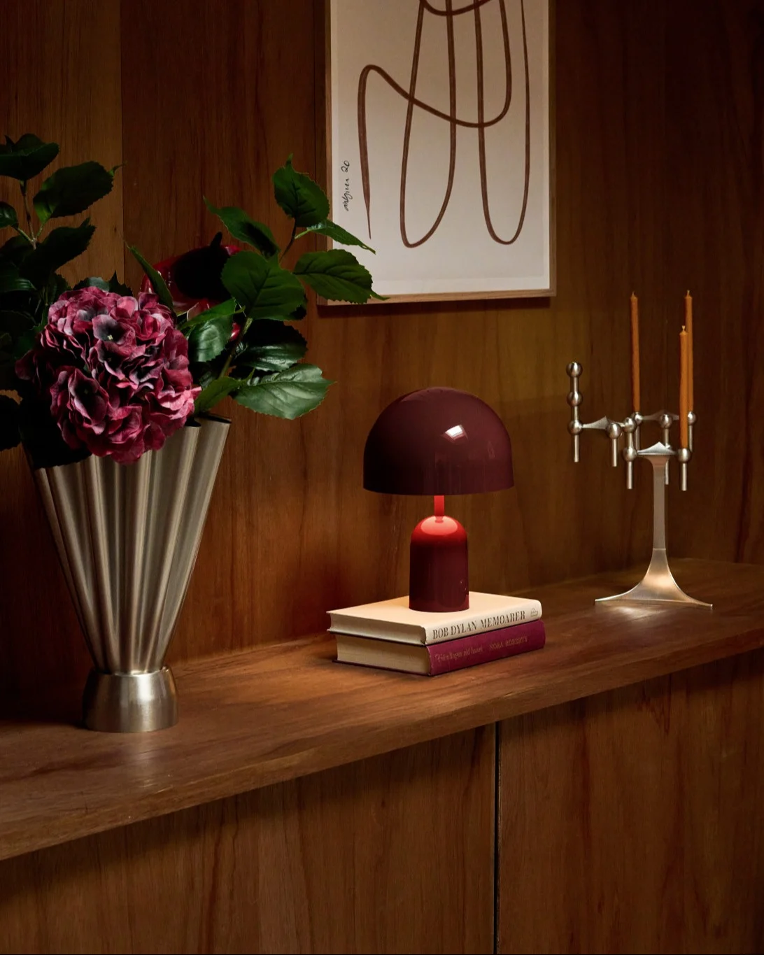 Mensola in legno con vaso argentato scanalato, ortensie rosse, lampada rossa su libri e candelabro argentato.