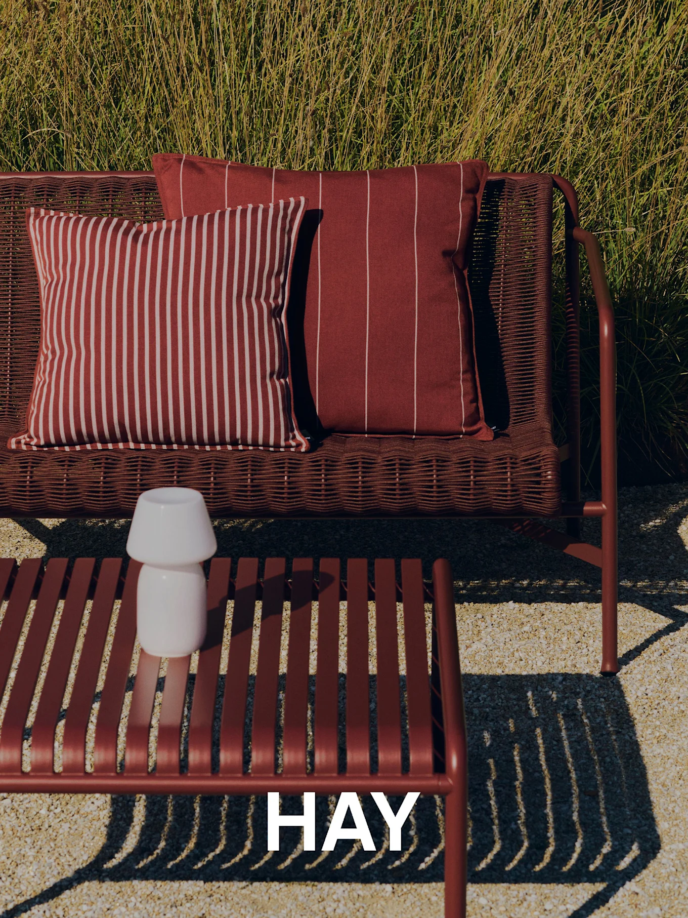 Scena esterna con un divano rosso intrecciato, cuscini a righe, un tavolino a doghe con lampada bianca ed erba alta sullo sfondo. Logo HAY.