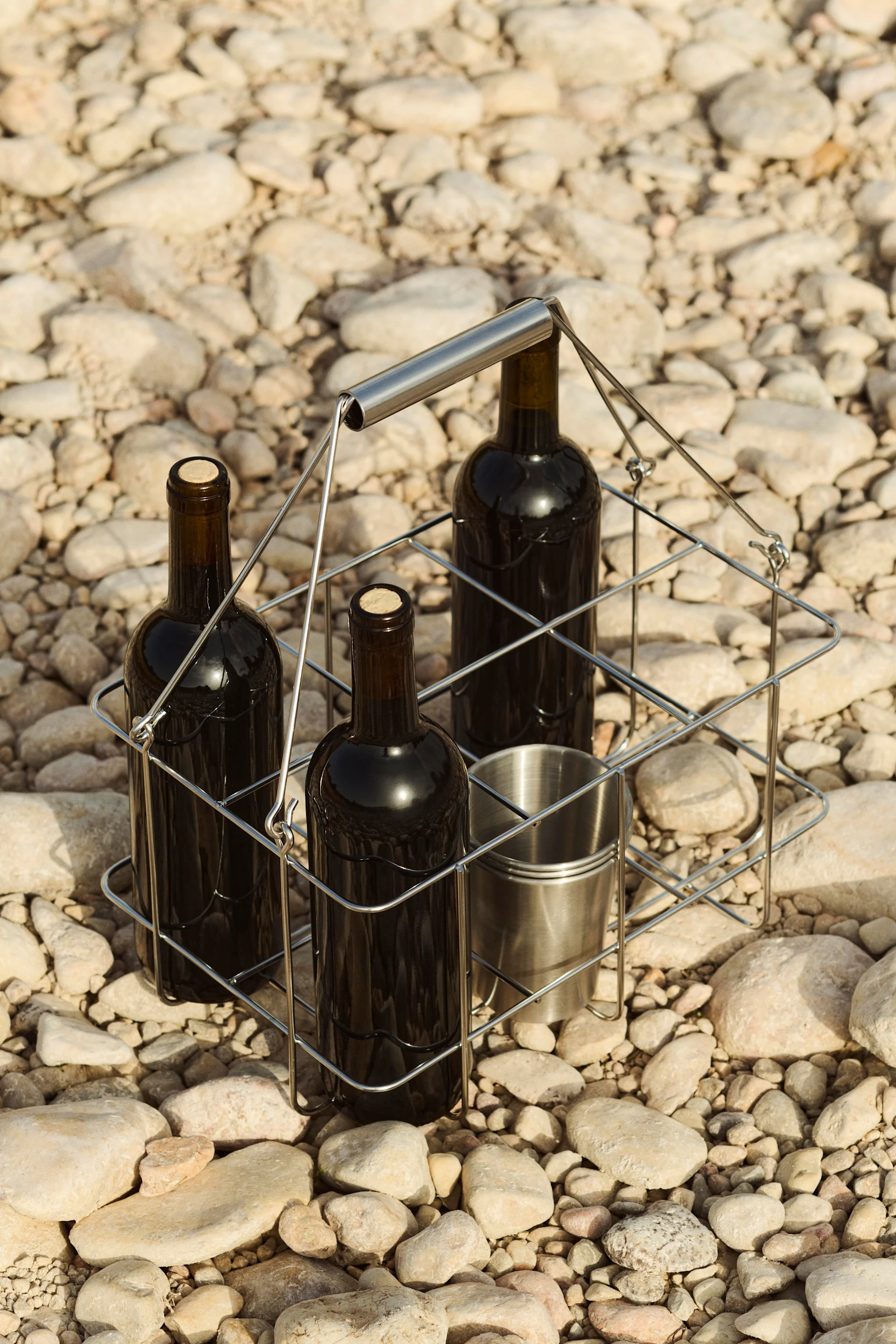 Tre bottiglie di vino e una tazza di metallo in un portabottiglie su un letto di ciottoli.