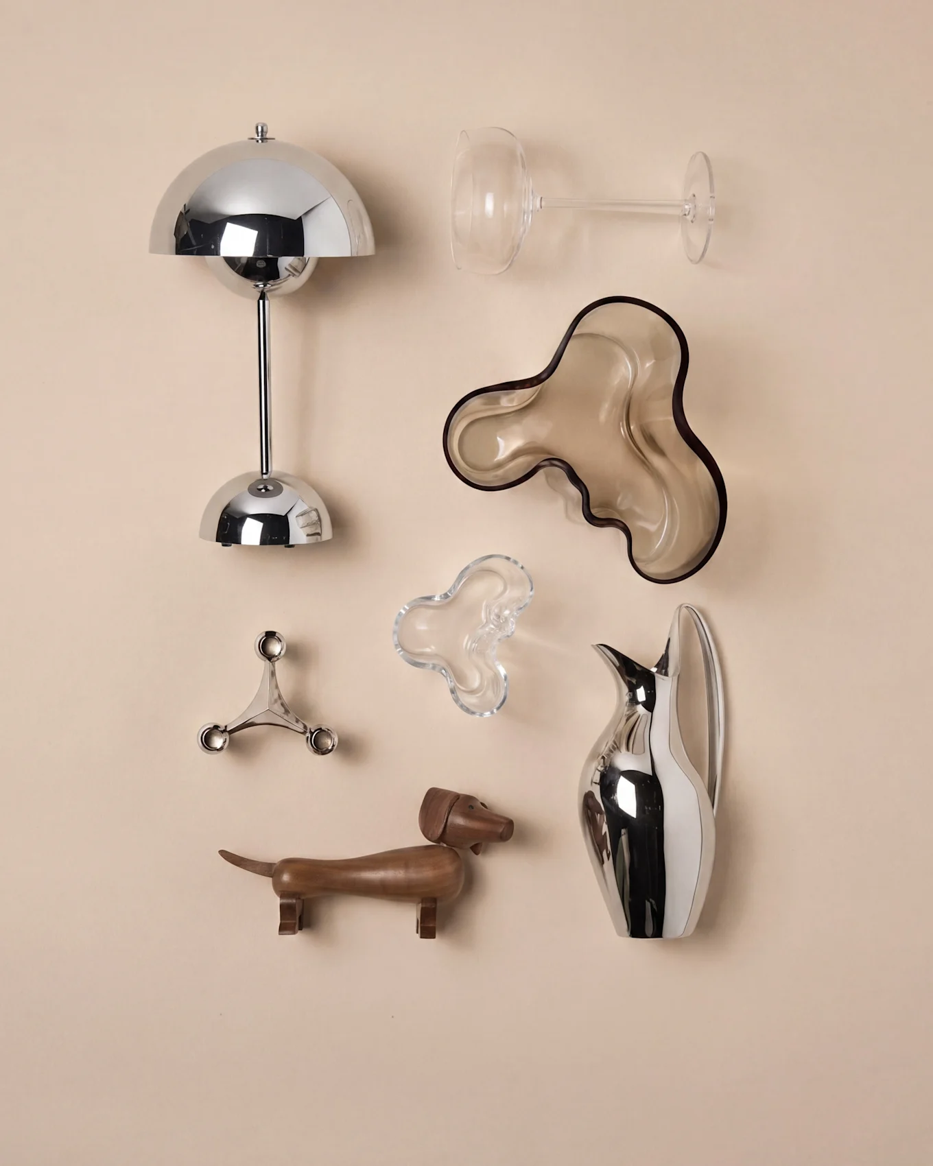 Una disposizione di oggetti decorativi: una lampada cromata, calice da vino, due ciotole organiche, un candelabro, un bassotto in legno e una brocca.