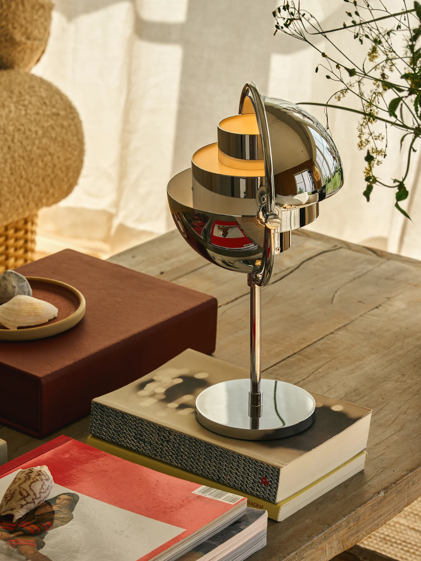 Lampada da tavolo cromata con paralumi sferici a più livelli che emettono un bagliore caldo, su un tavolo di legno con libri e conchiglie.