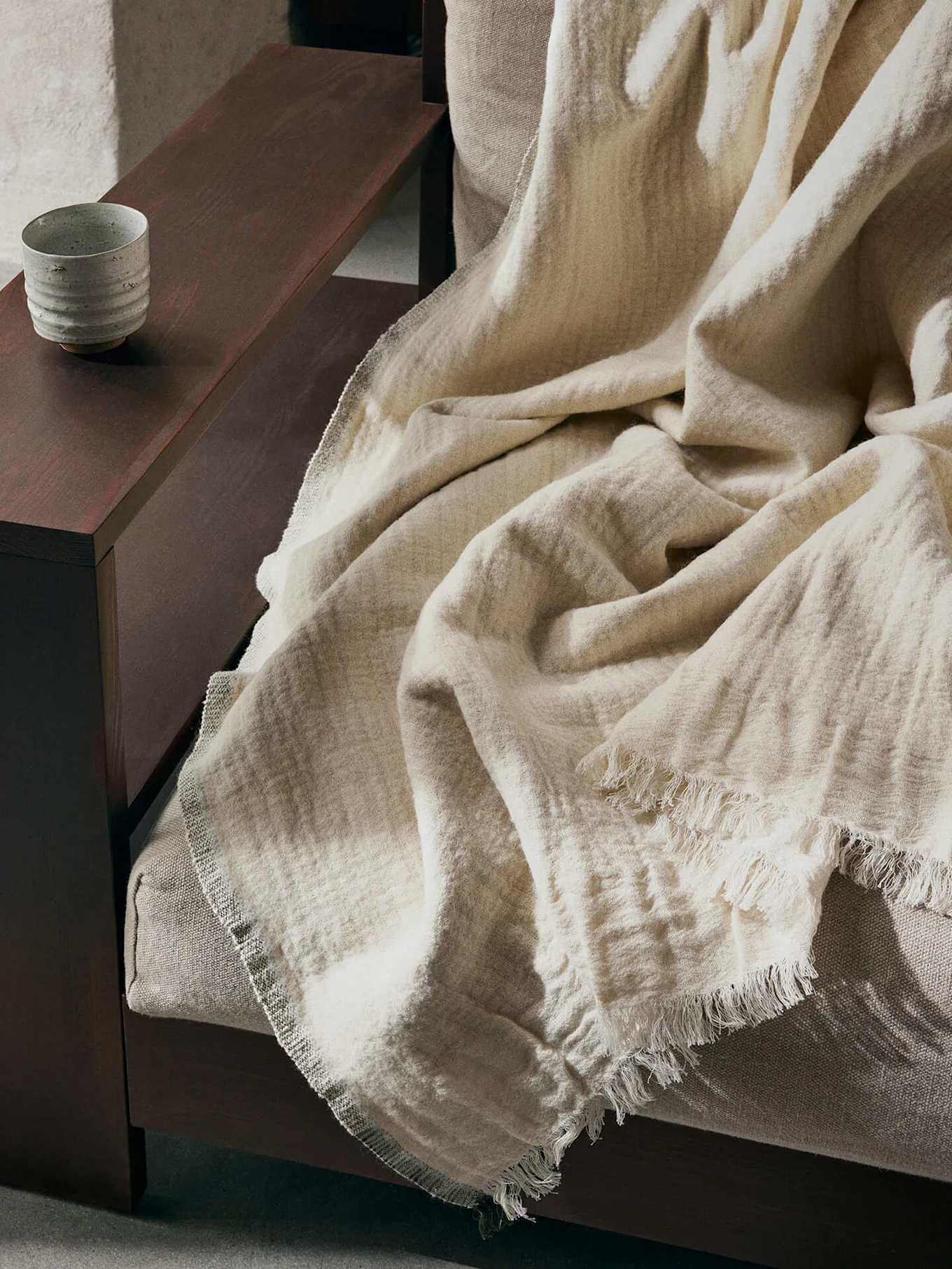 Una coperta testurizzata beige chiaro con bordi sfrangiati drappeggiata su un divano, con una tazza di ceramica su un bracciolo di legno scuro.