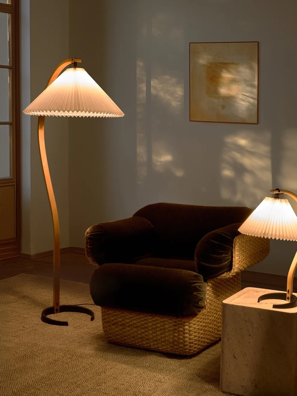 Stanza accogliente con lampada da terra curva, poltrona scura con base intrecciata e lampada da tavolo più piccola. La luce calda crea ombre.