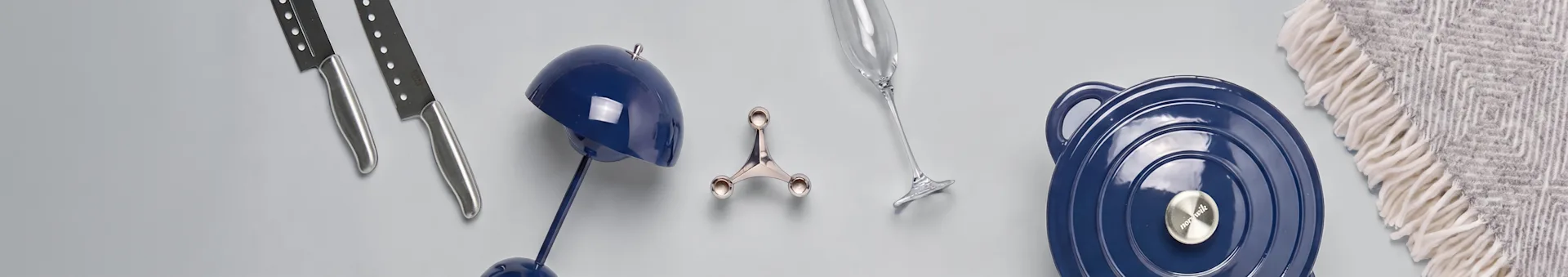Due coltelli, una lampada blu, un supporto argentato, un flûte da champagne, una pentola blu e una coperta grigia a fantasia su una superficie chiara.