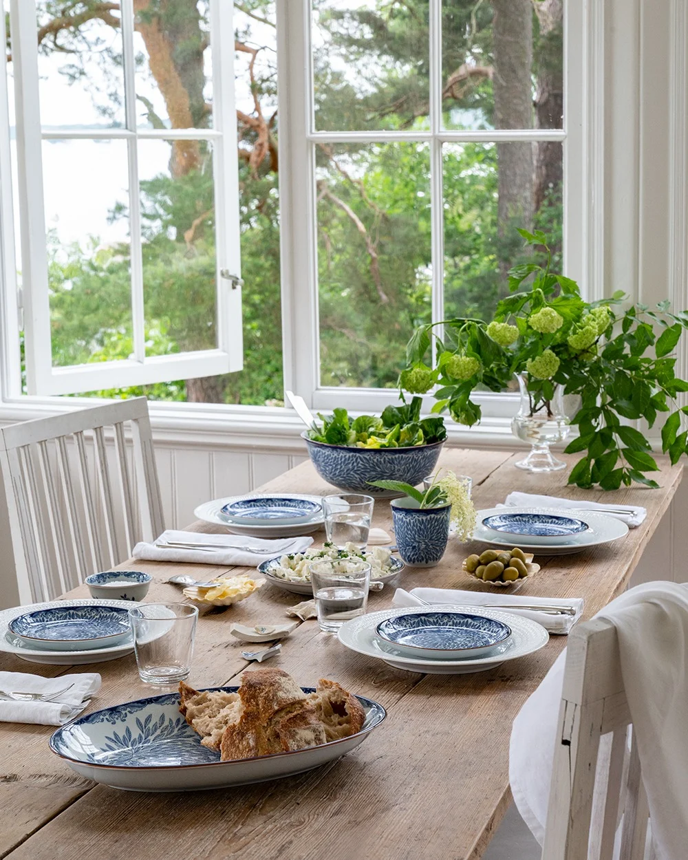Un tavolo da pranzo rustico in legno apparecchiato con piatti blu e bianchi, insalata, pane e olive, accanto a una finestra aperta che affaccia su alberi verdi e acqua.