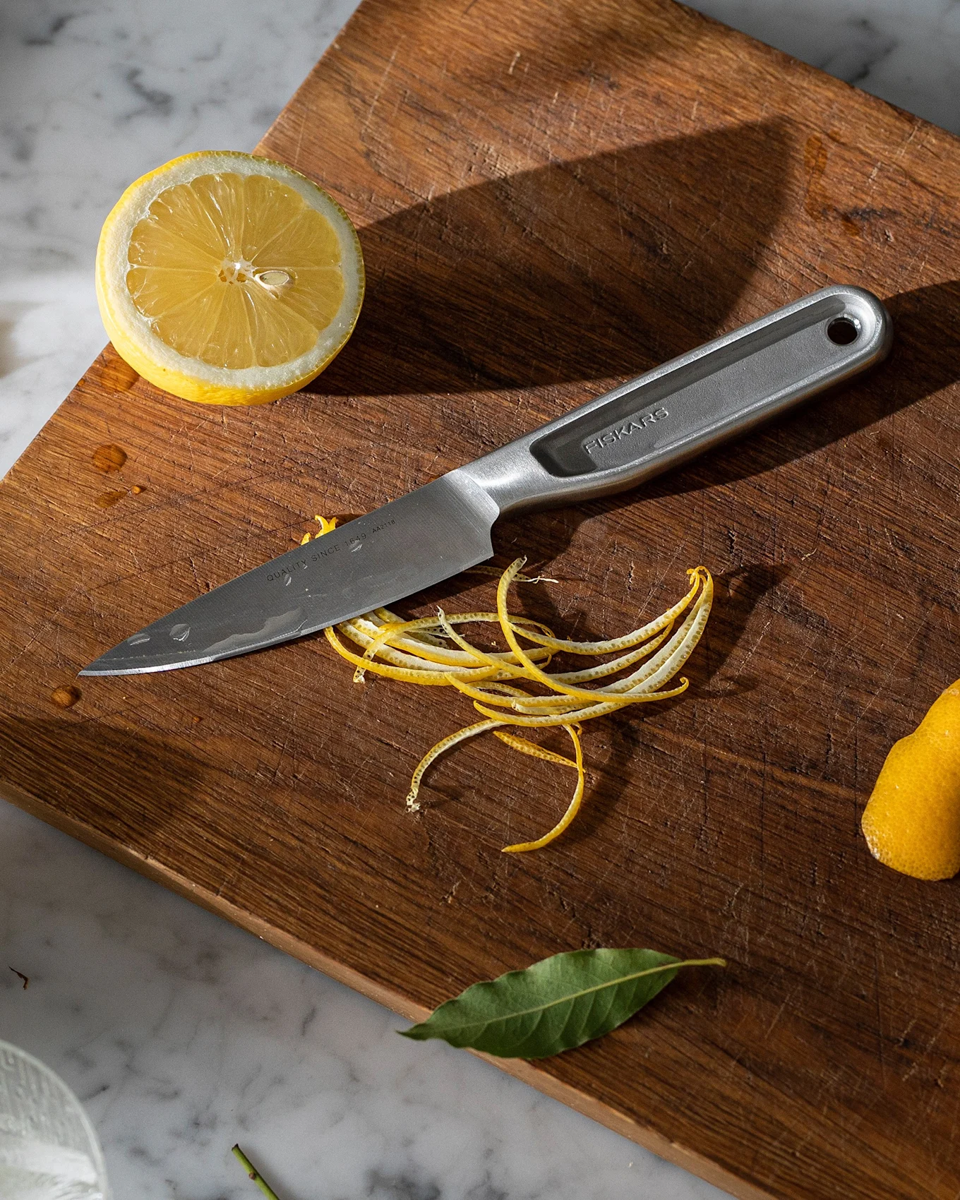 Un coltello in acciaio inossidabile su un tagliere di legno con mezzi limoni, bucce di limone e foglie di alloro su un bancone di marmo con un bicchiere di ghiaccio.