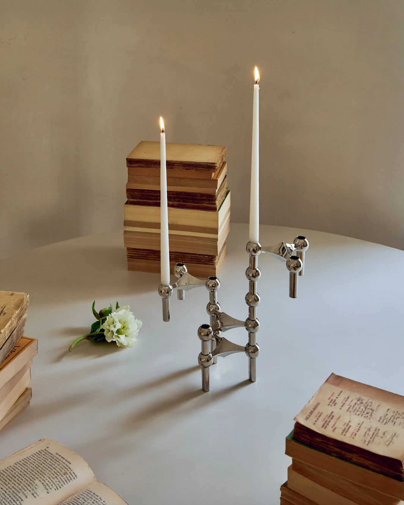 Due portacandele modulari cromati con candele bianche accese, circondati da vecchi libri e un fiore bianco su un tavolo.