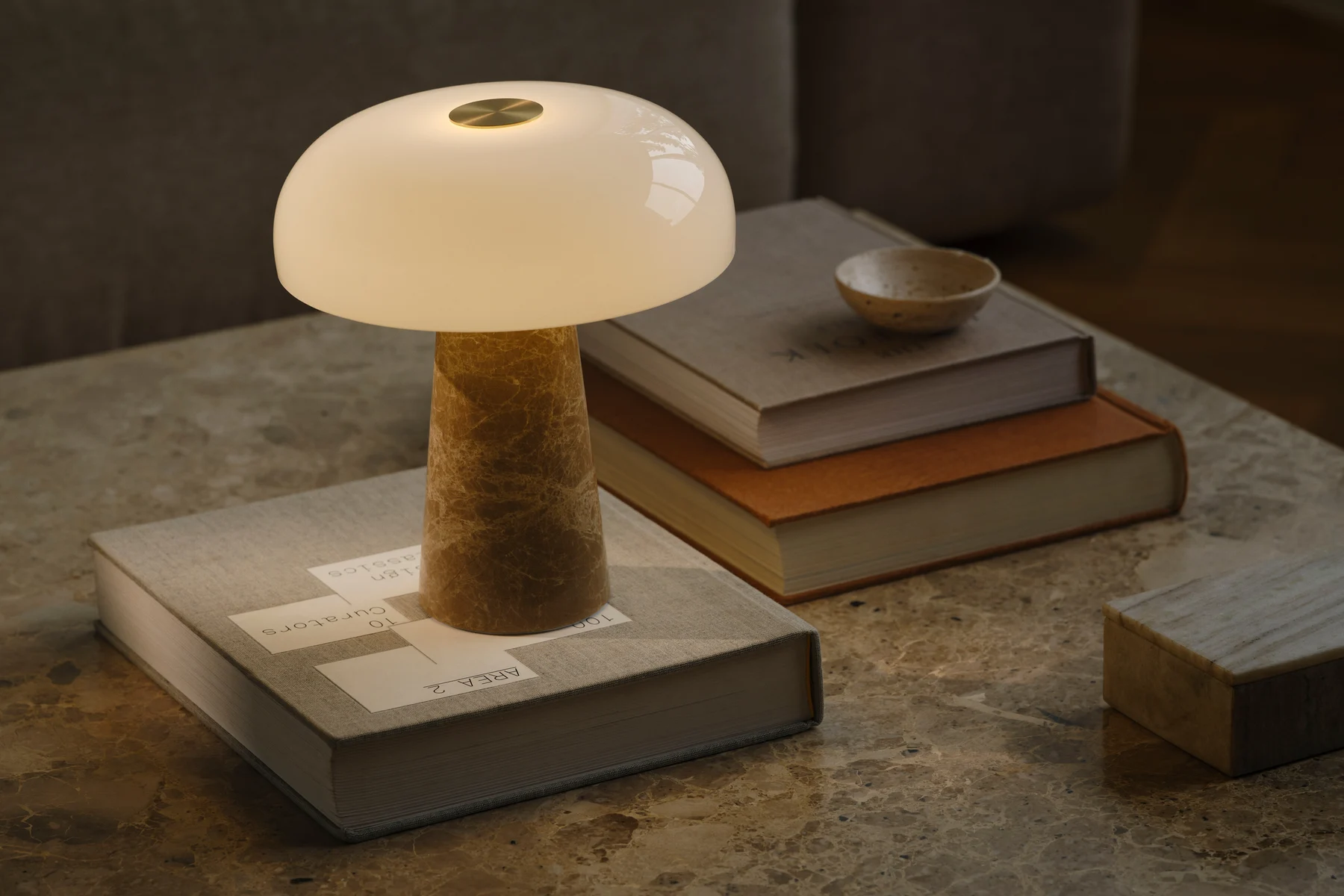 Una lampada da tavolo a forma di fungo con paralume in vetro bianco e base conica in marmo, su un tavolo di marmo con libri impilati.
