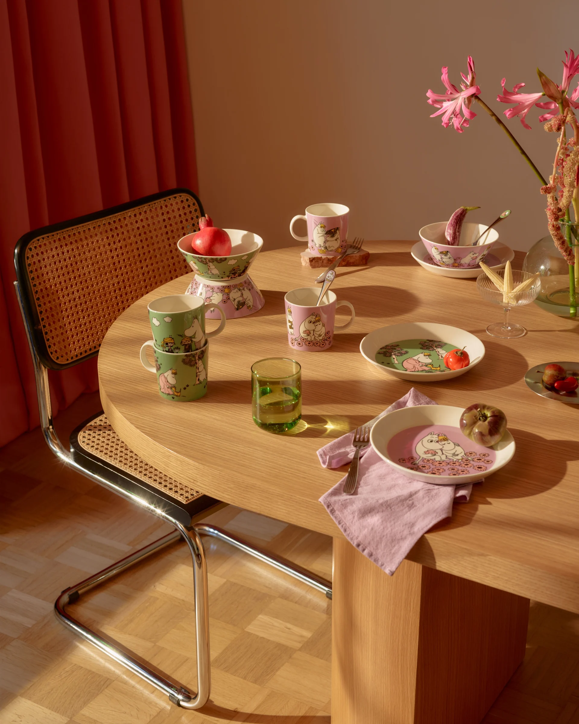 Tavolo da pranzo con stoviglie a tema Moomin rosa e verdi, frutta fresca, un bicchiere verde e fiori rosa.