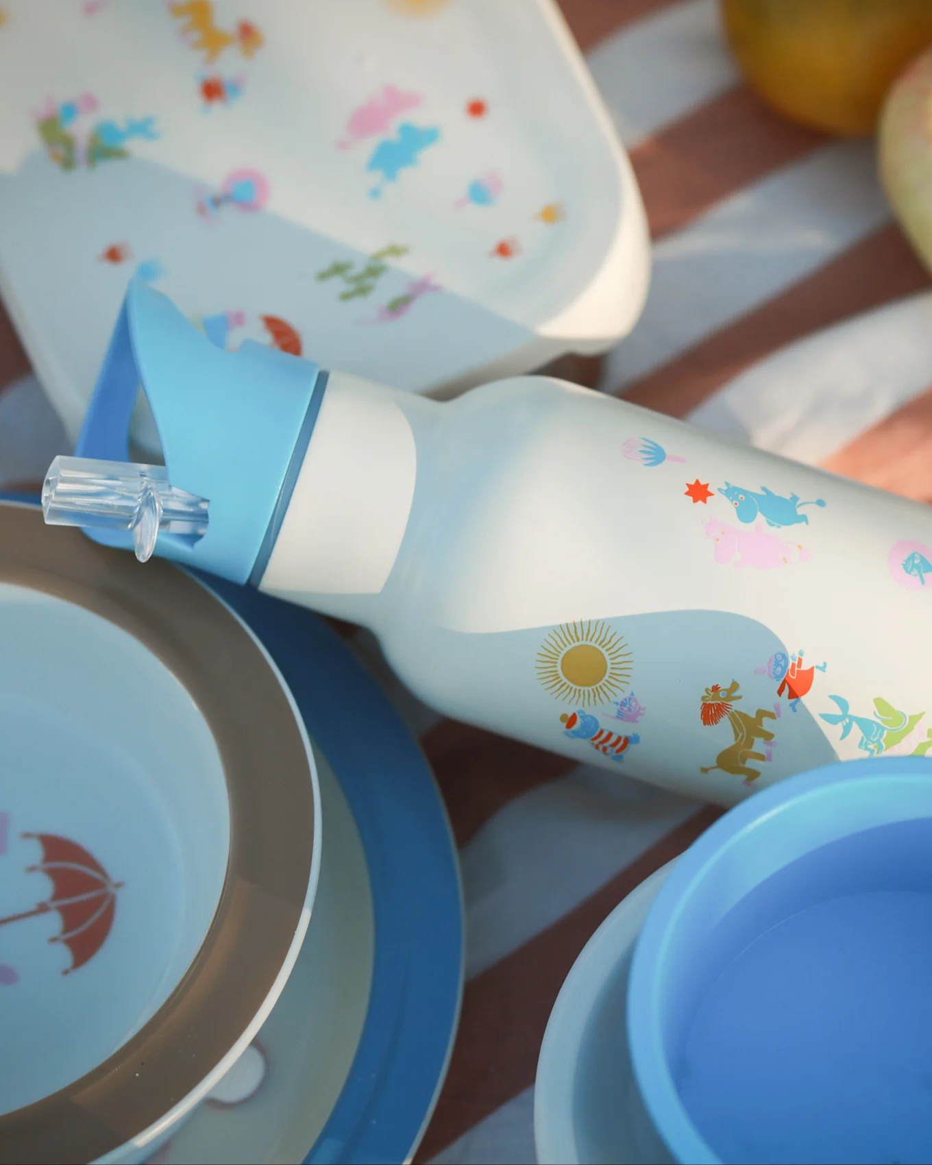 Borraccia bianca con cannuccia blu, personaggi dei cartoni animati colorati e sole, circondata da piatti da picnic abbinati su una coperta a righe.