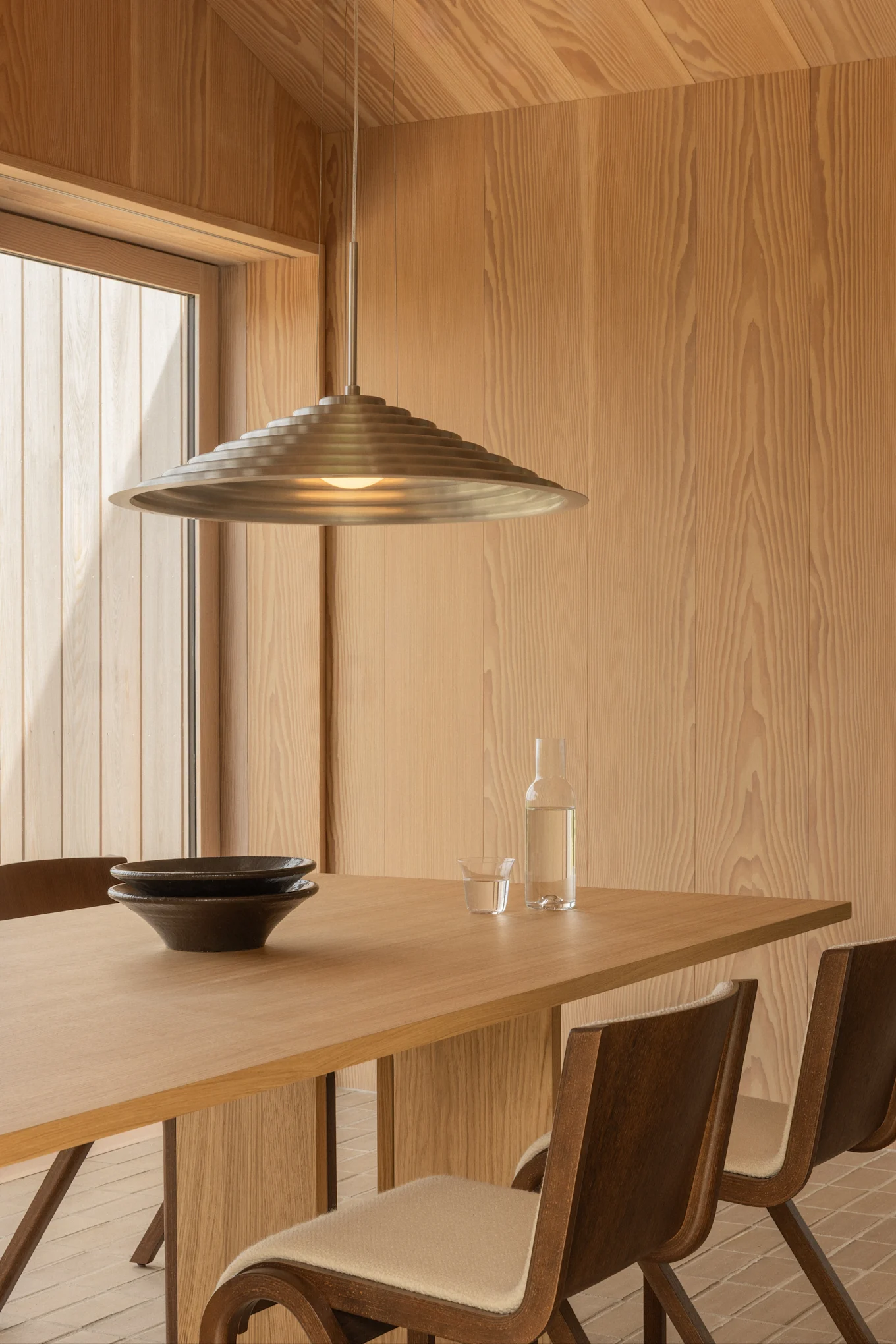 Sala da pranzo moderna con pareti in legno chiaro, un tavolo in legno con due ciotole scure, una bottiglia d'acqua e una lampada a sospensione a più livelli in metallo.