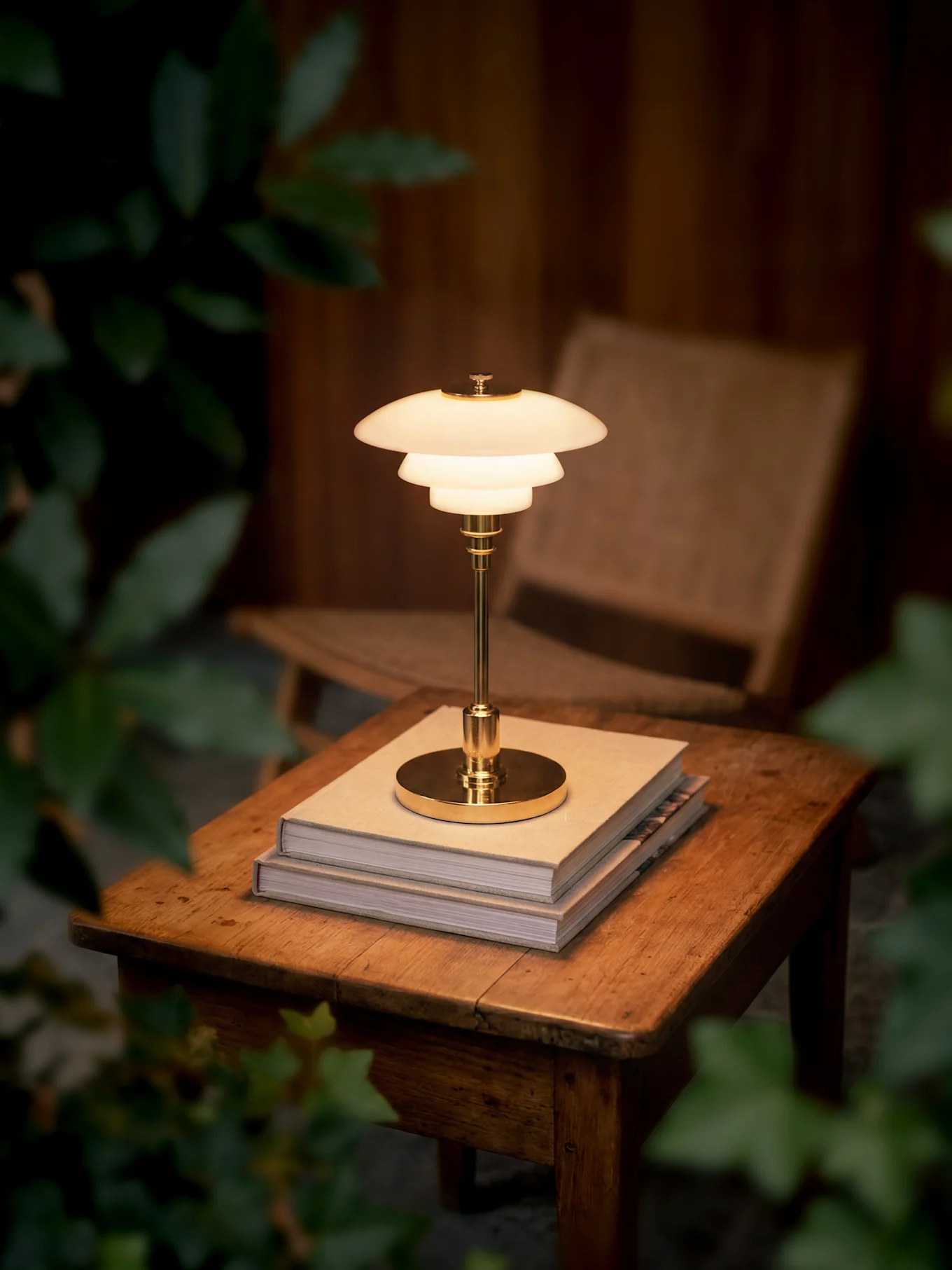 Una lampada da tavolo in ottone lucida a più livelli poggia su due libri su un tavolo di legno, circondata da fogliame scuro.