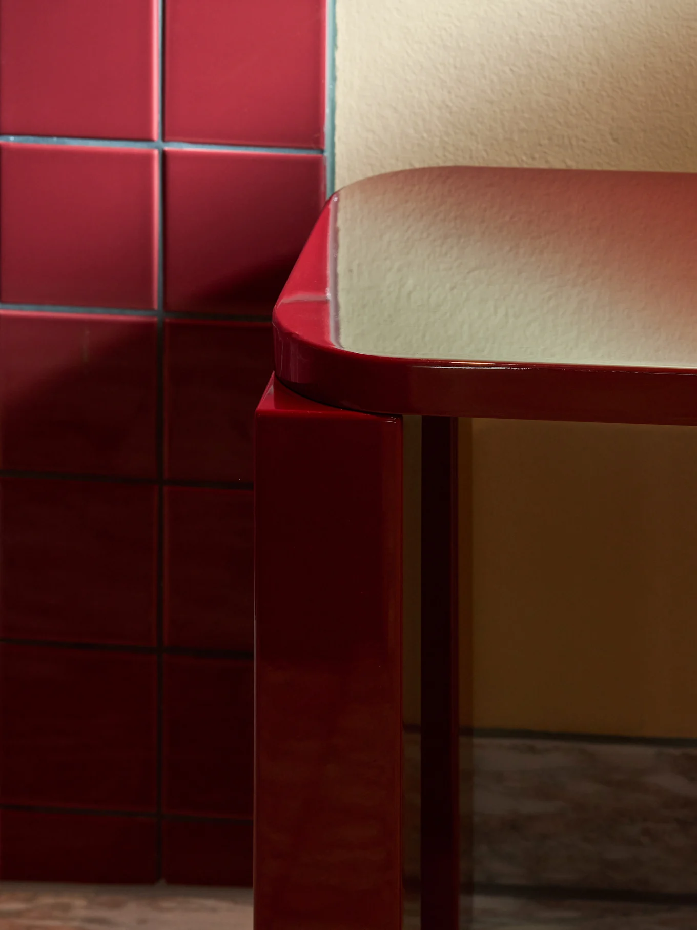 Elegante e lucido sgabello rettangolare rosso con bordi arrotondati su pavimento in marmo, accanto a parete di piastrelle rosse.