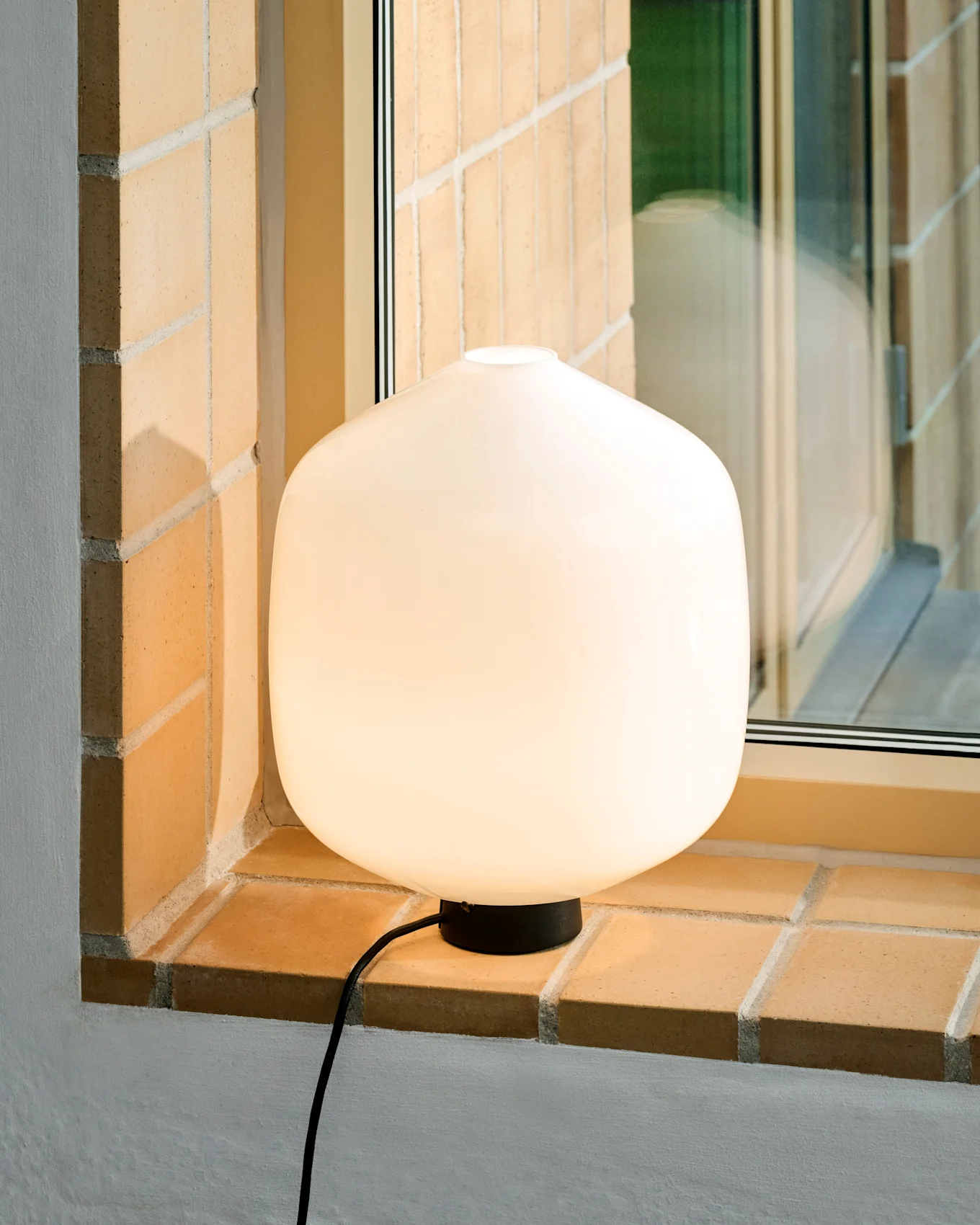 Una lampada da tavolo rotonda bianca e luminosa con base e cavo neri è posizionata su un davanzale di mattoni chiari.