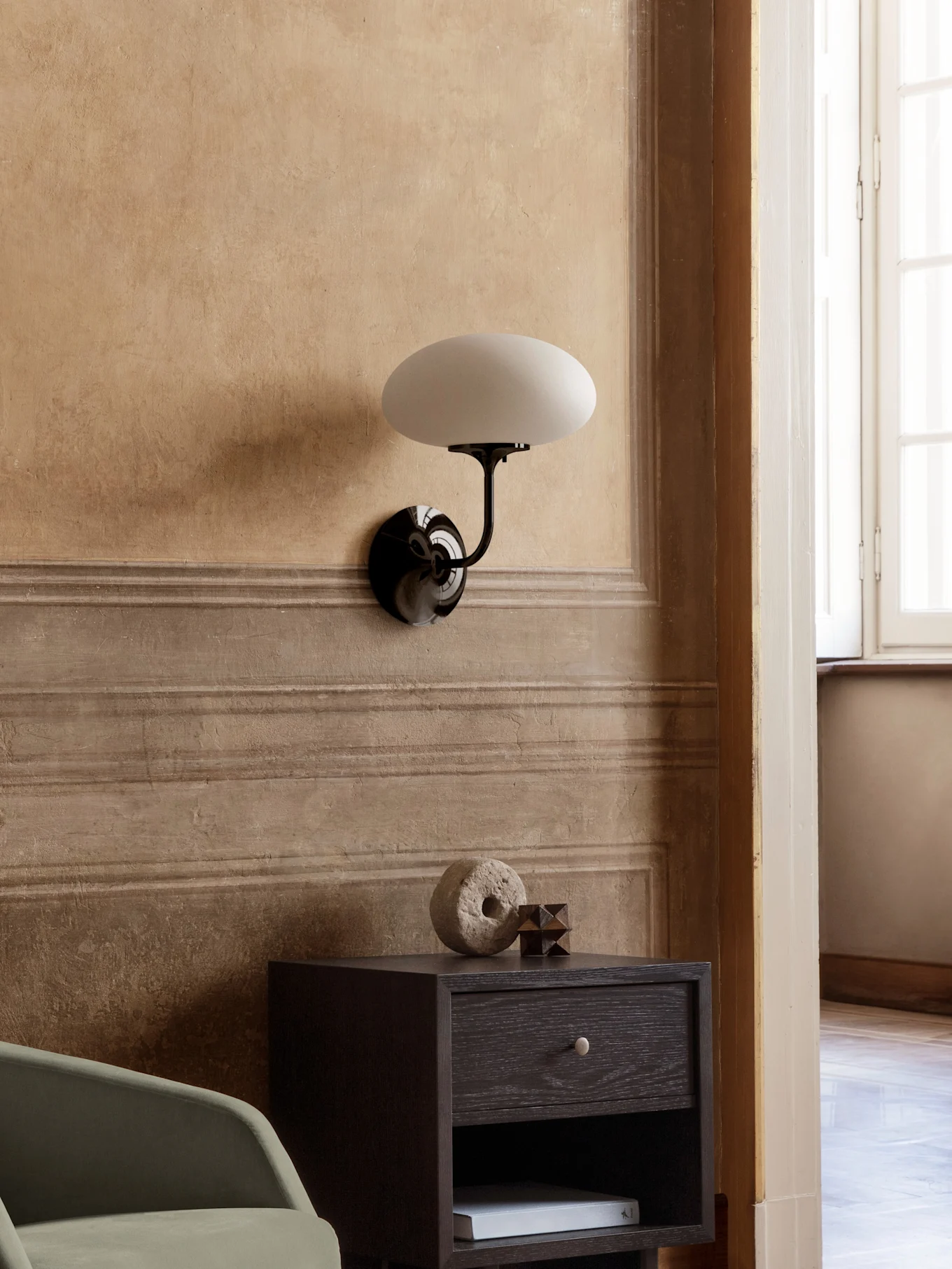 Lampada da parete con braccio nero e paralume ovale bianco sopra comodino in legno scuro con decori.