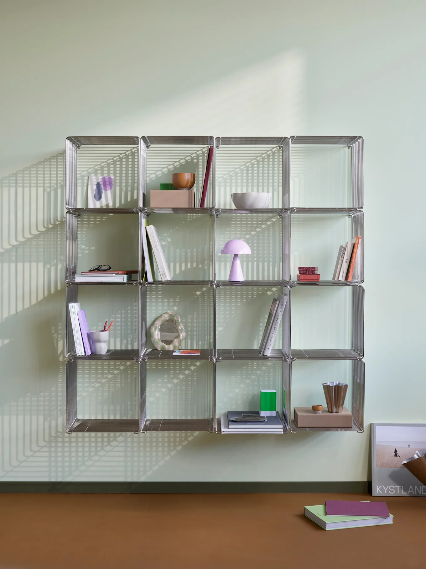 Una libreria modulare color cromo con libri e oggetti decorativi, illuminata da forti ombre lineari su una parete verde chiaro e pavimento marrone.