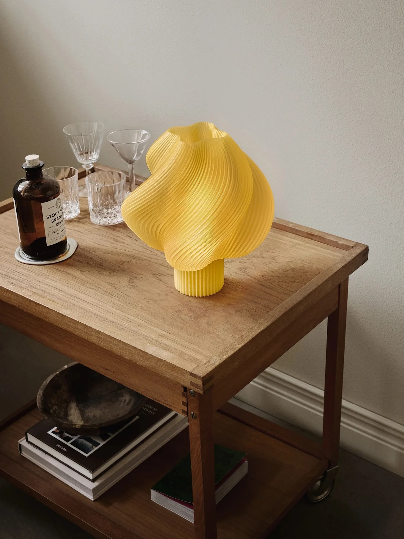 Lampada gialla plissettata su carrello in legno con bottiglia di gin, bicchieri e libri sullo scaffale inferiore.